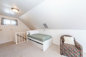 A small, single-bed room with a sloped ceiling, a window with curtains, a radiator, and a patterned armchair.