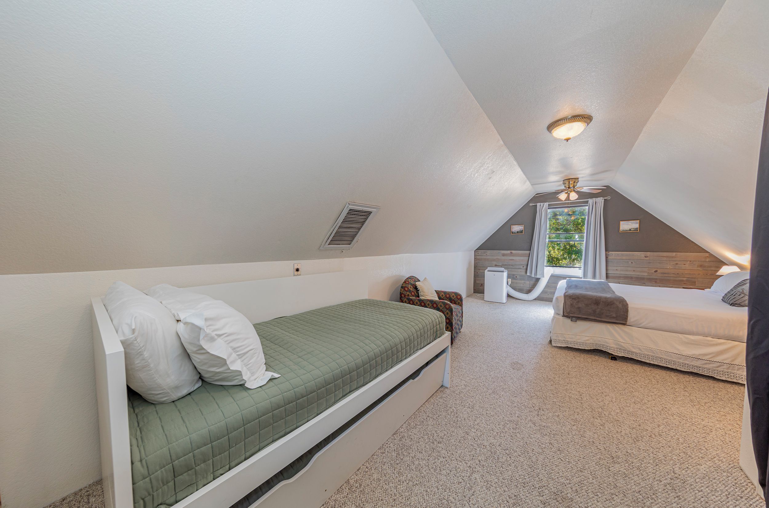 A small, sloped ceiling room with a bed, a chair, a heater, and a window with curtains.