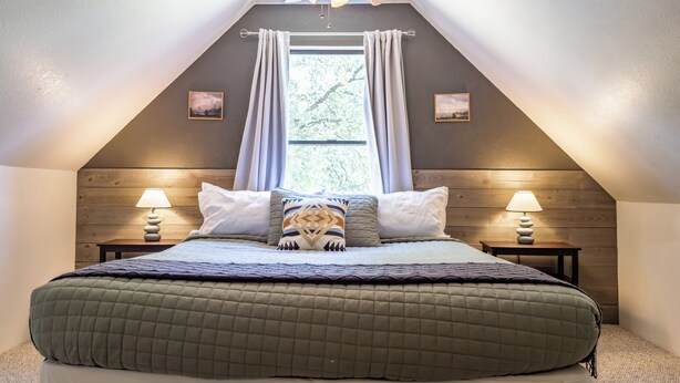 Un dormitorio con techo inclinado, una cama con colcha verde acolchada, dos lámparas de noche y un ventilador de techo.