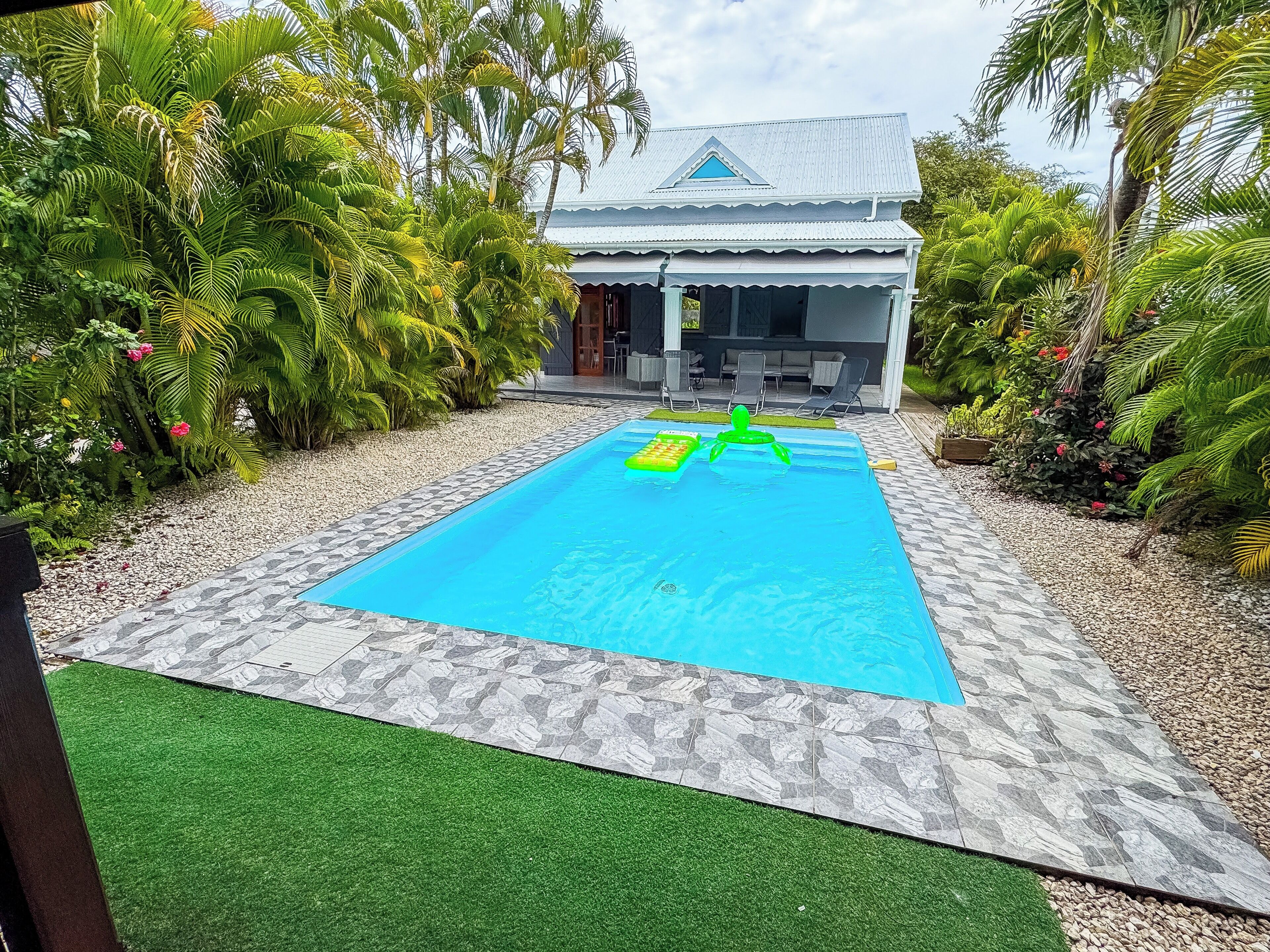 Uma piscina com duas espreguiçadeiras insufláveis, rodeada por plantas tropicais e uma casa com telhado azul.