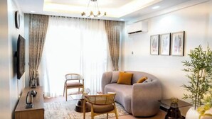 Ein modernes Wohnzimmer mit einer Couch, einem Esstisch und Stühlen. Ein großes Fenster mit Vorhängen, eine Kristallleuchter und gerahmte Kunstwerke an der Wand sind zu sehen.