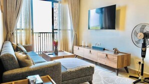 Ein modernes Wohnzimmer mit Sofa, Hocker, Couchtisch und einem an der Wand befestigten Fernseher. Es gibt einen Holzschrank und einen Standventilator.