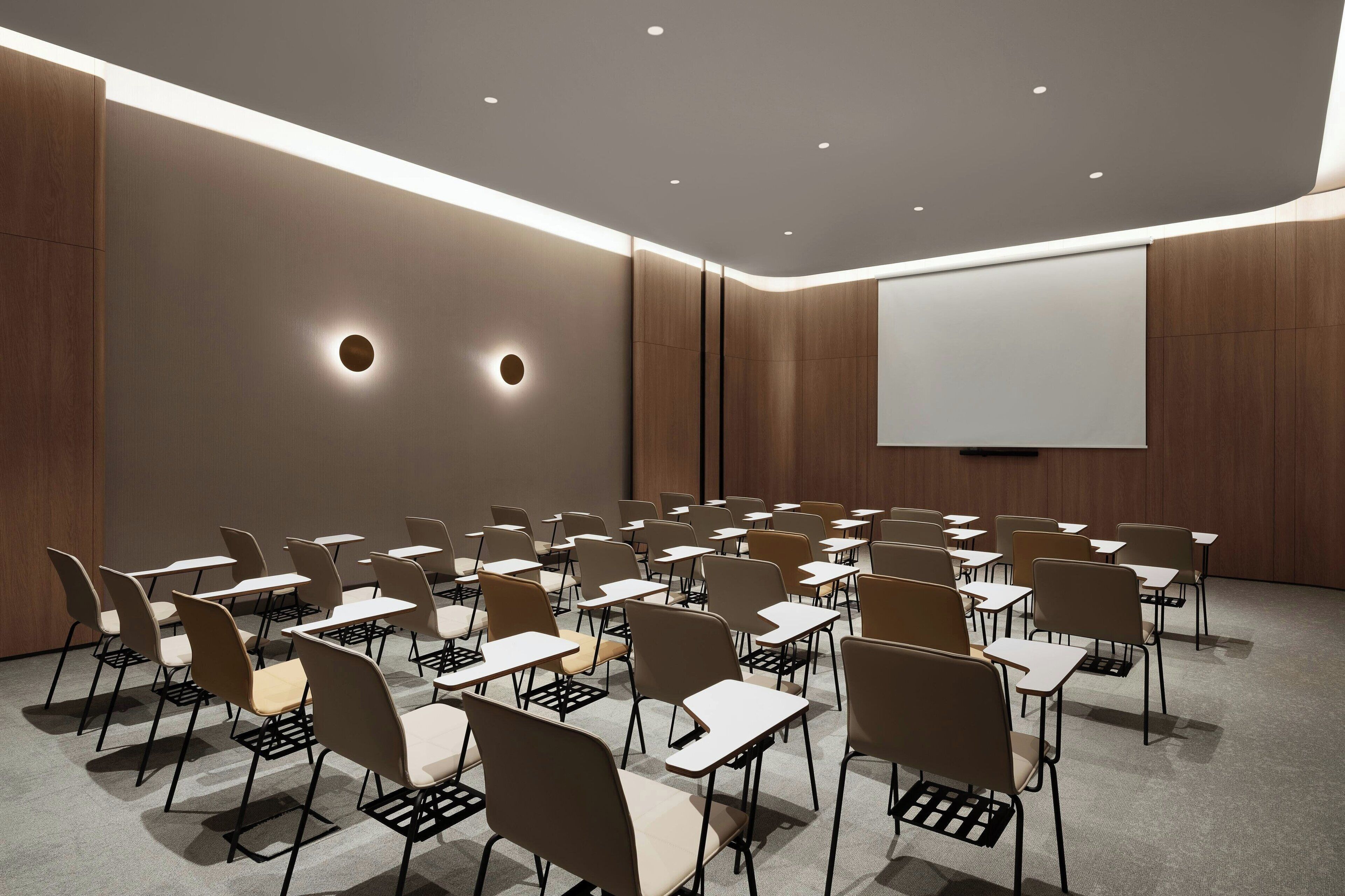 A conference room with rows of chairs and tables, a projector screen, and recessed ceiling lighting.