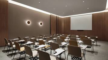 A conference room with rows of chairs and tables, a projector screen, and recessed ceiling lighting.
