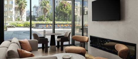 A modern living room with a sofa, chairs, a coffee table, and a fireplace. A TV is mounted on the wall, and there is a view of buildings and trees outside.
