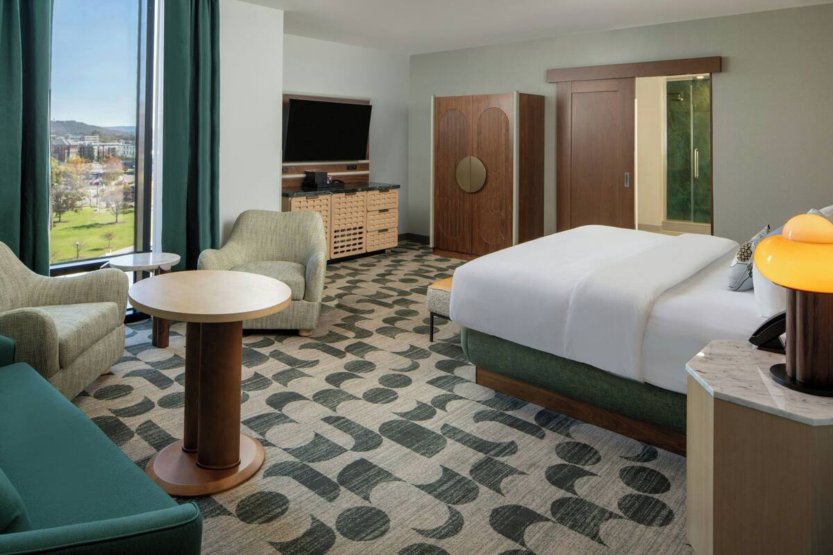 A hotel room with a bed, two armchairs, a small table, a TV, a wardrobe, and a view of the outdoors.