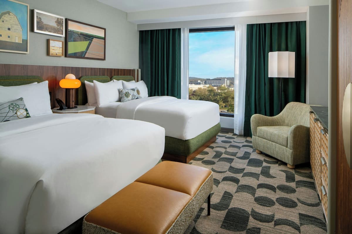 A hotel room with two beds, a sofa, a bench, a lamp, and a view of the city.