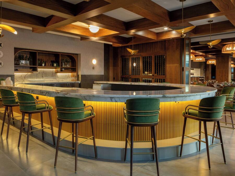 A bar area with a curved counter, green bar stools, wooden beams, and pendant lighting.
