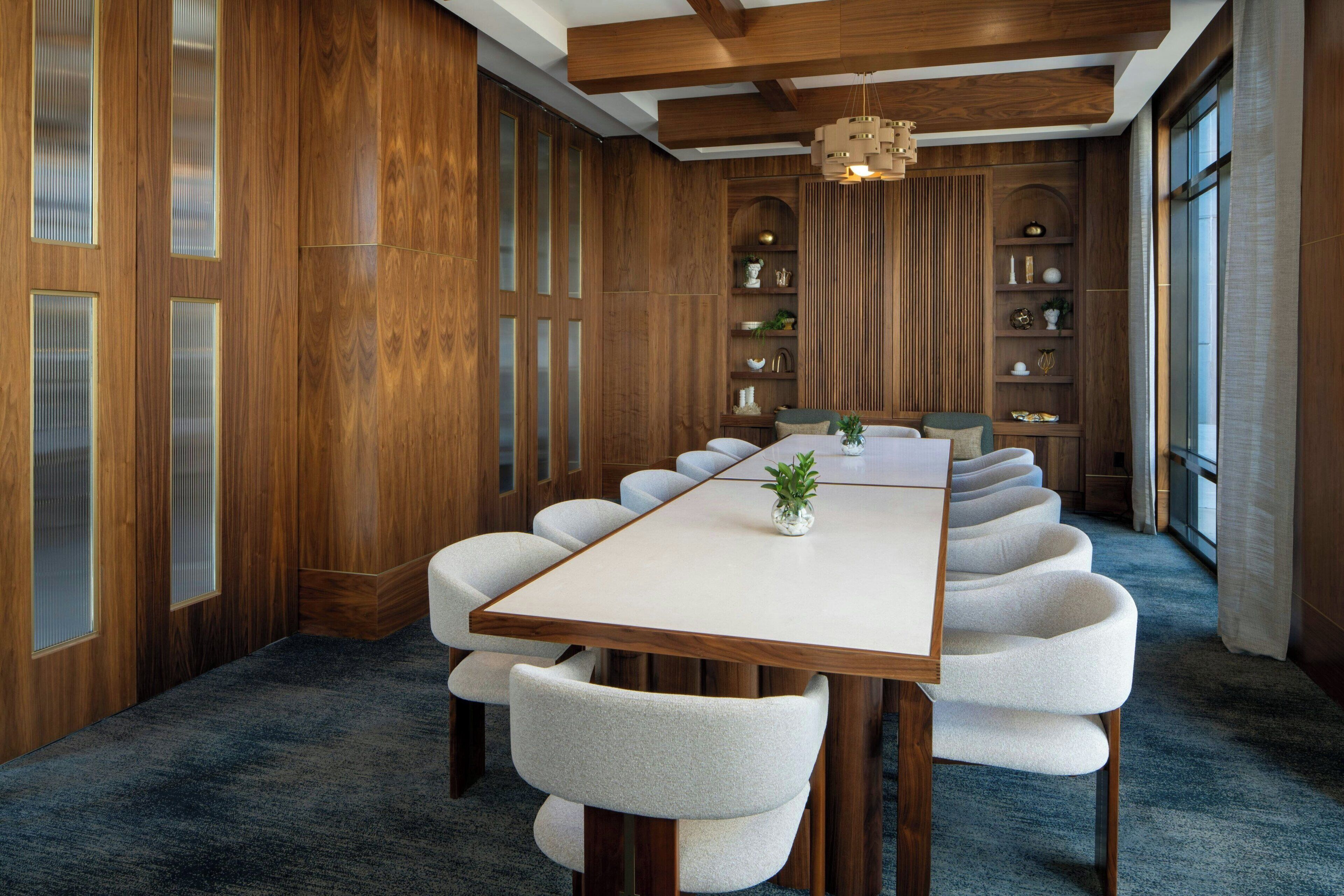 Une salle de conférence avec une longue table, des chaises blanches rembourrées, un lambris en bois et une plante décorative.