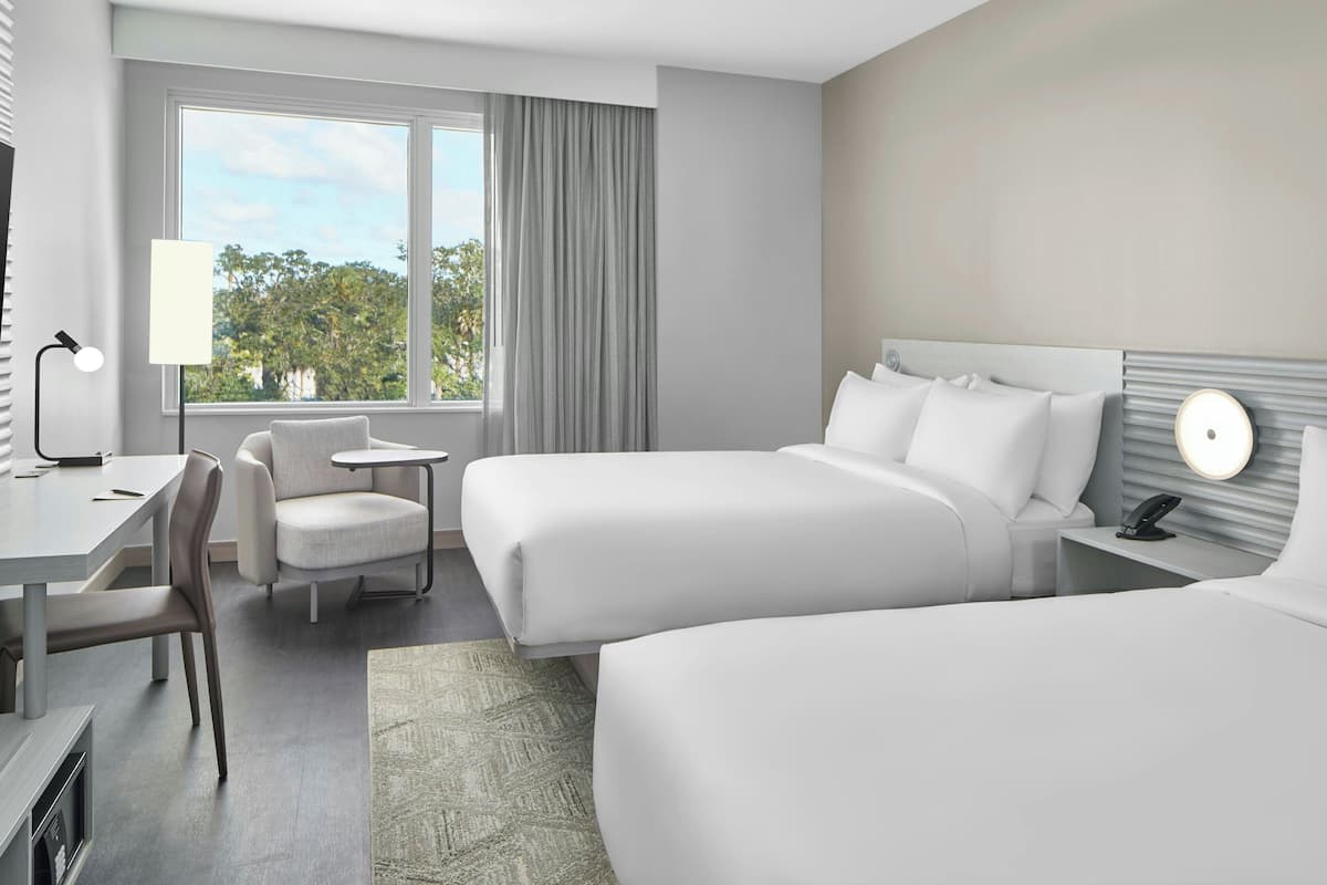 A hotel room with two beds, a desk, a chair, a lamp, and a view of trees through the window.