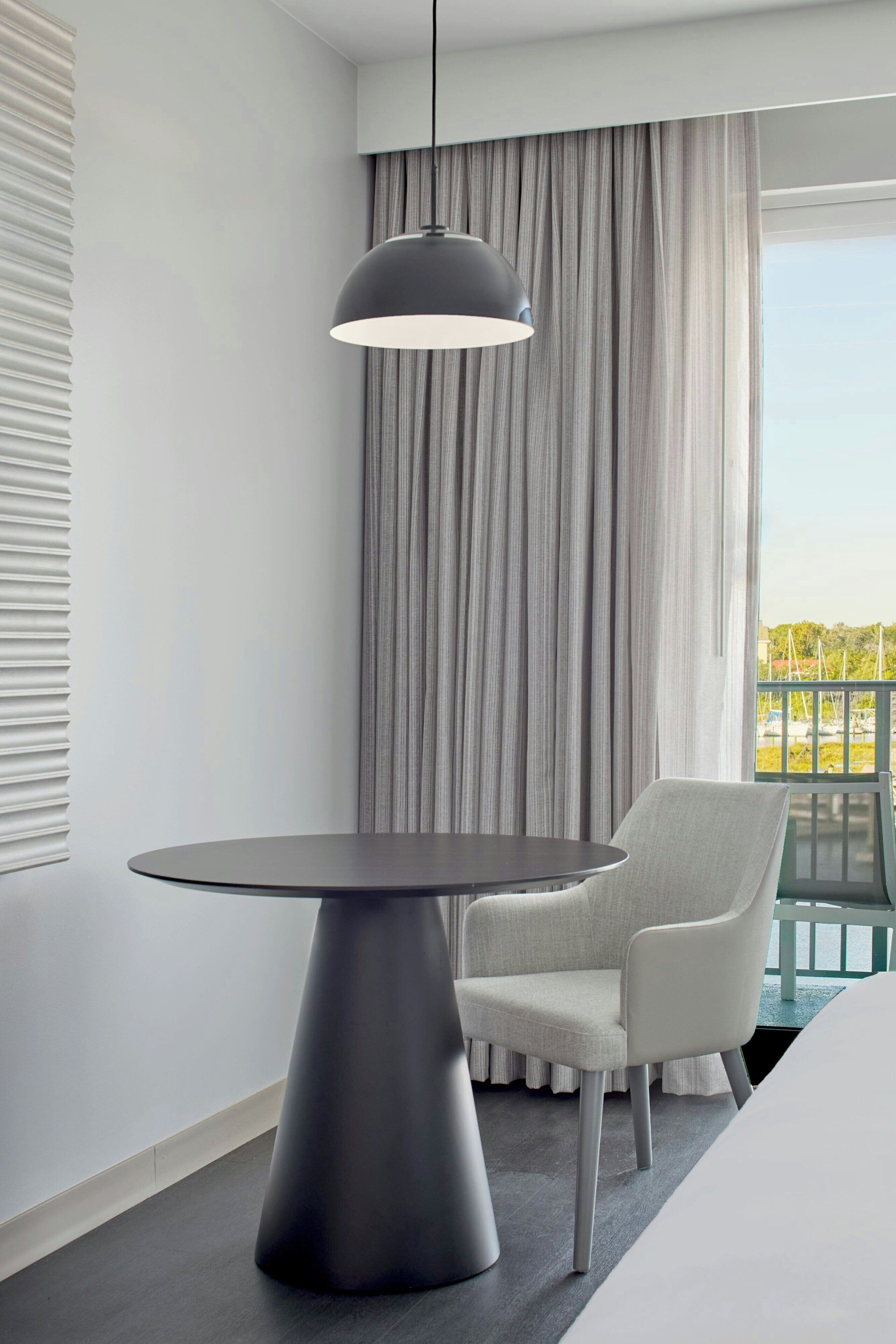 A modern room with a round table, a chair, a pendant light, and a view of the outdoors through a window with curtains.