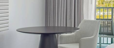 A modern room with a round table, a chair, a pendant light, and a view of the outdoors through a window with curtains.