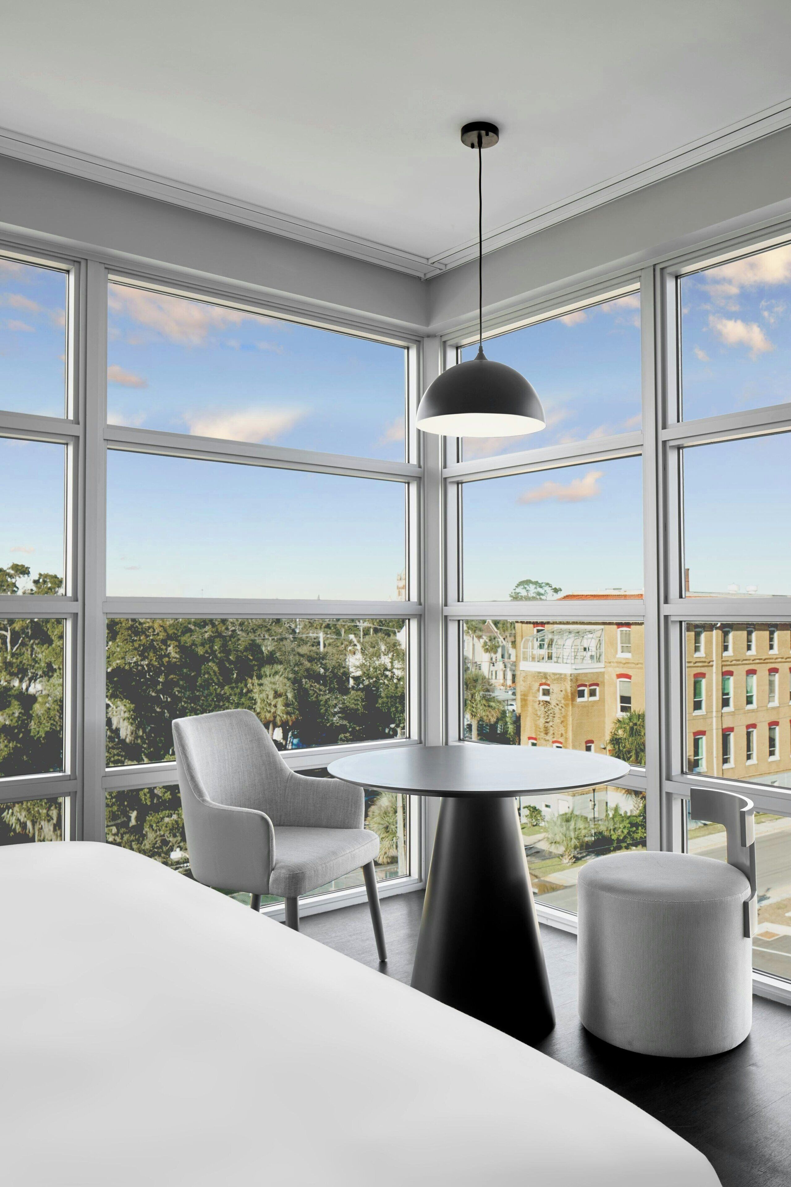 A modern hotel room with large windows, a round table, a chair, and a hanging light fixture.