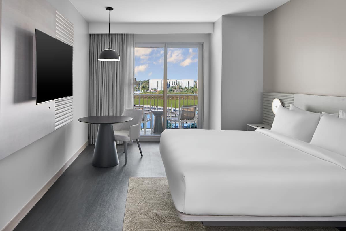 A modern hotel room with a large bed, a flat-screen TV mounted on the wall, a small round table with chairs, and a balcony with a view of buildings and greenery.