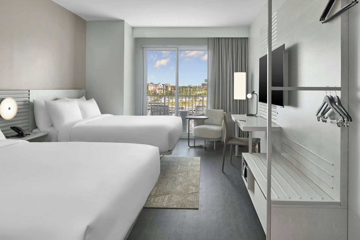 A modern hotel room with two beds, a desk, a chair, a TV, and a view of the city.