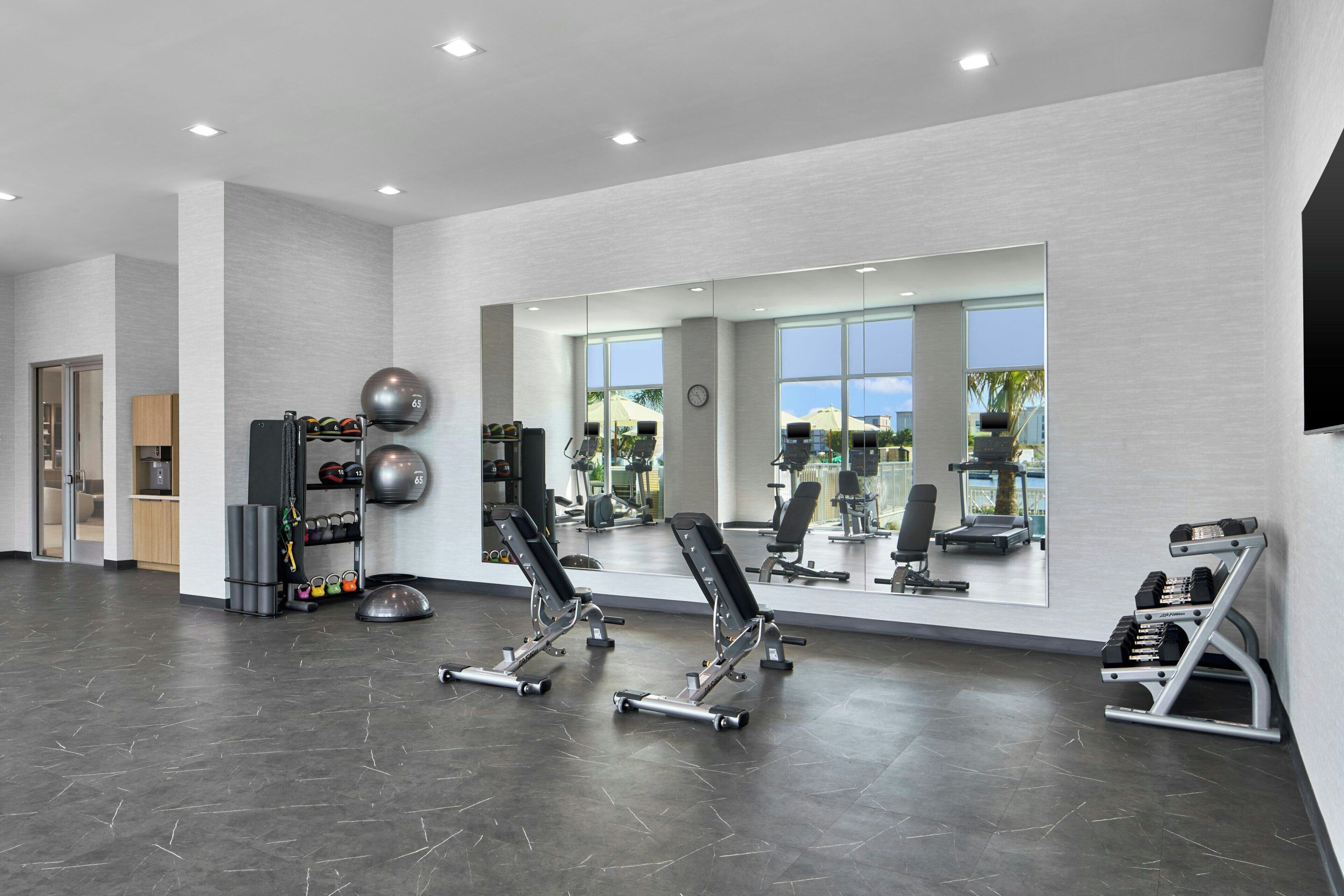 A modern gym with exercise equipment, large windows, and a mirror reflecting the equipment.