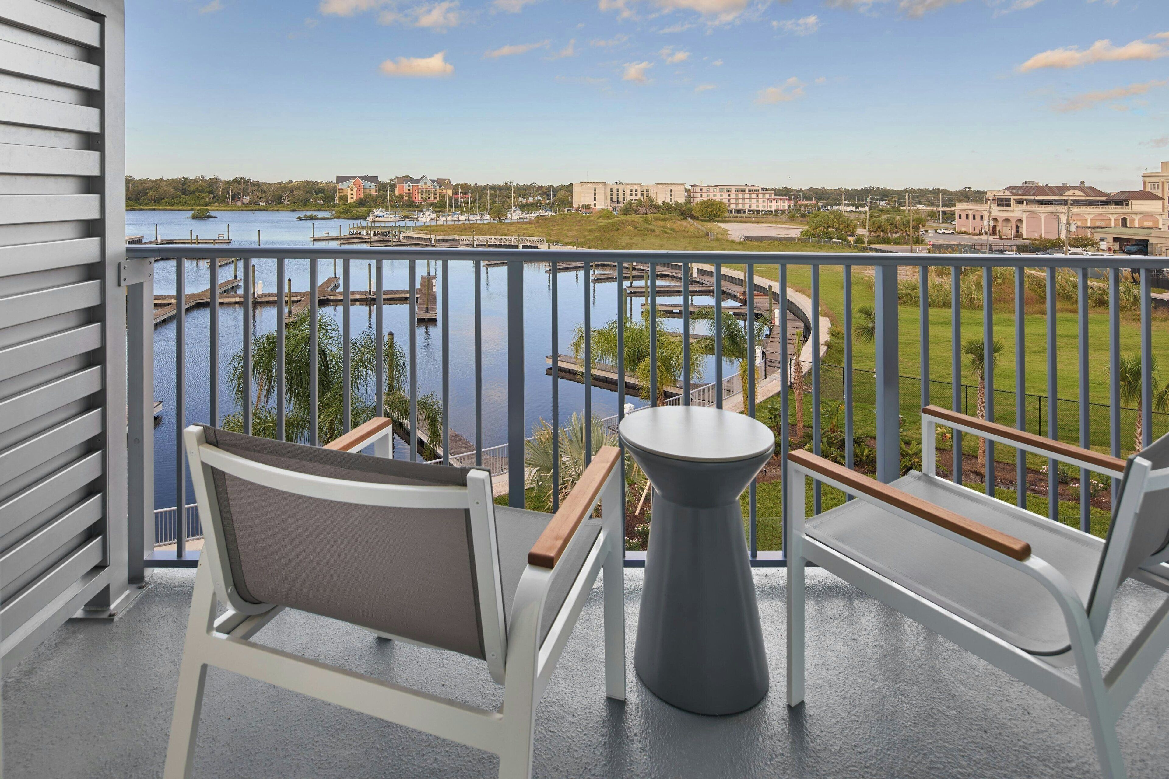 En balkon med to hvide stole og et gråt bord, med udsigt over en lystbådehavn med både og en græspl æne med bygninger i baggrunden.