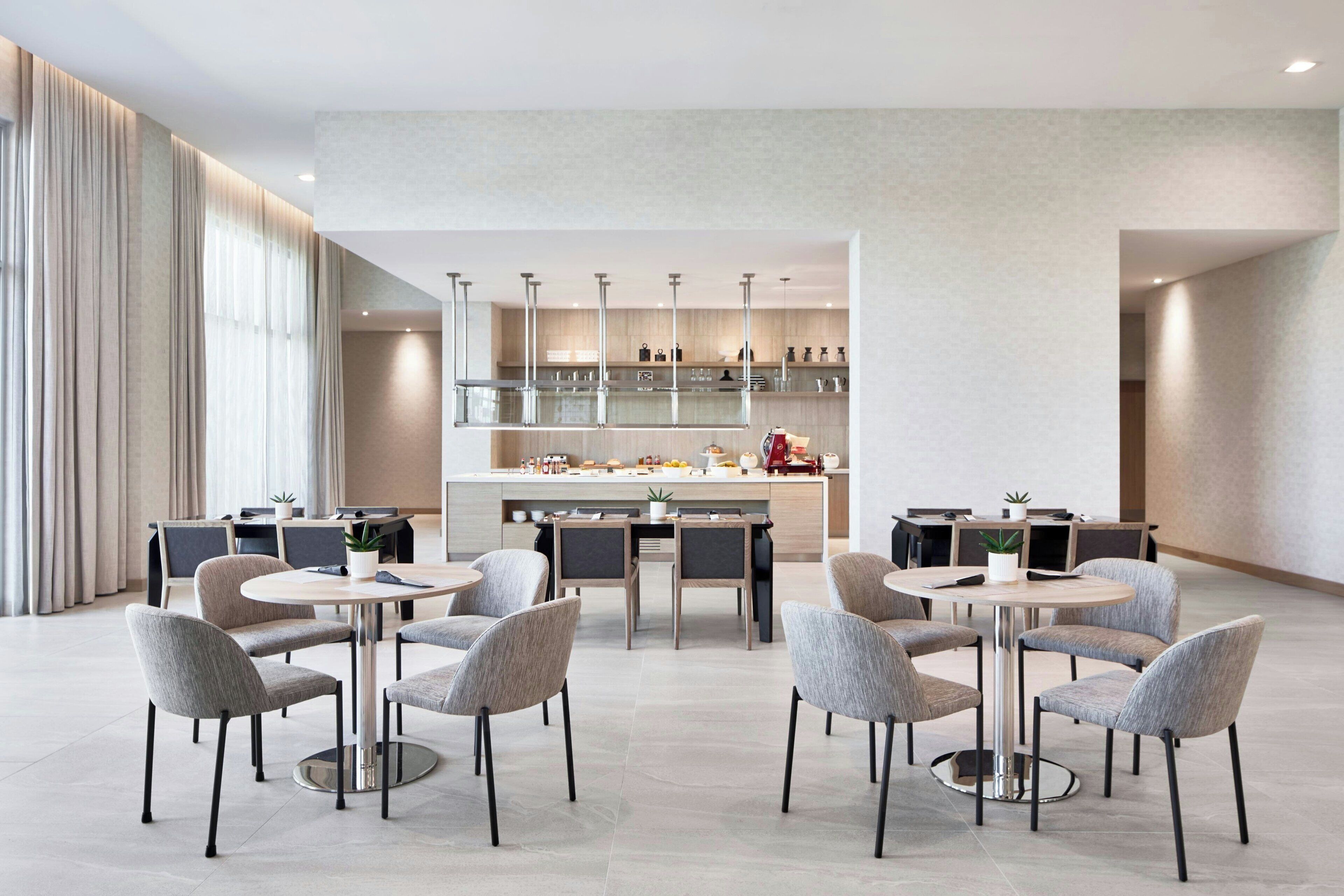 A modern dining area with round tables and chairs, a kitchen in the background, and a large window with curtains.