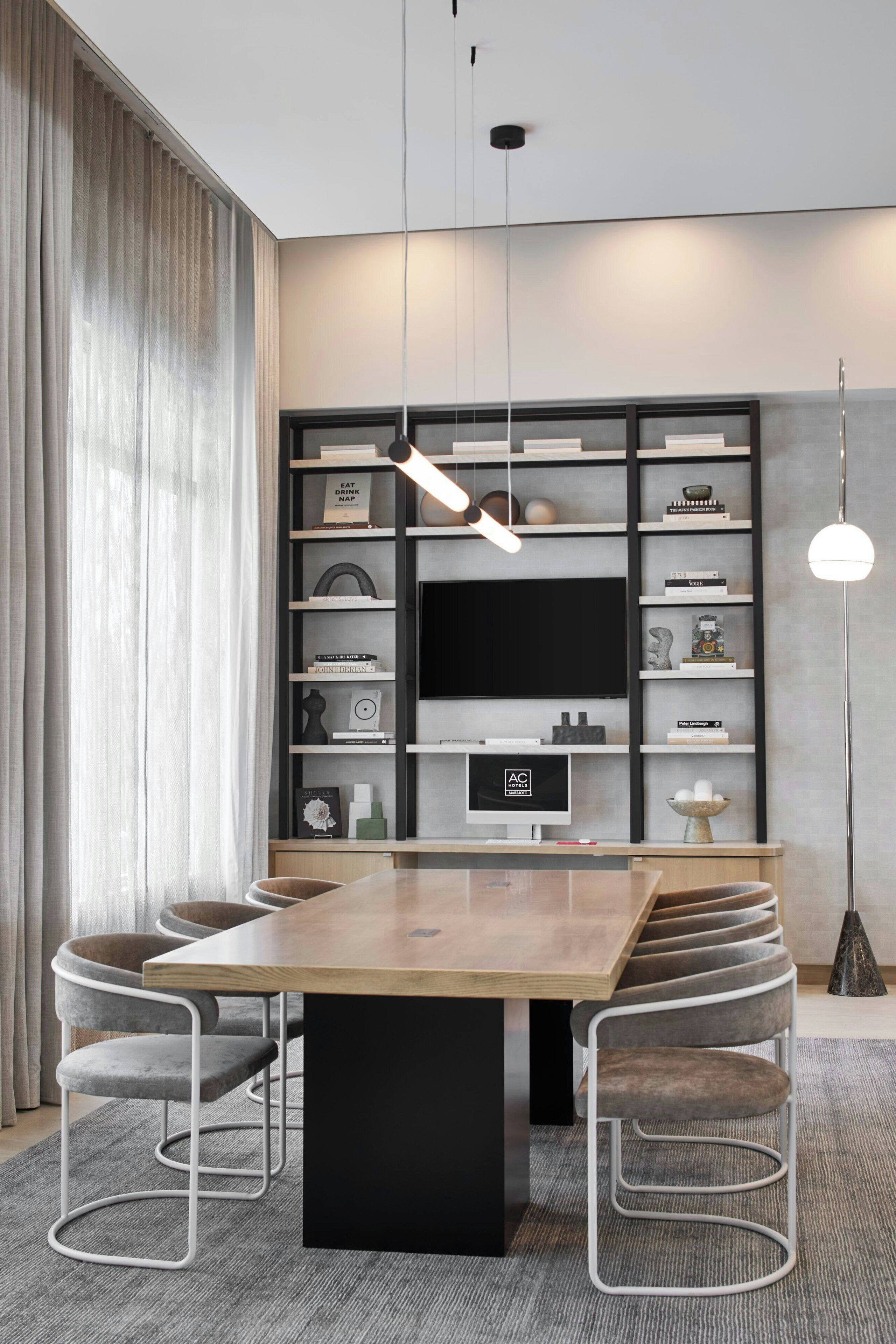 Un espace repas moderne avec une longue table en bois, des chaises à revêtement gris et une console centrale noire. Un grand téléviseur à écran plat est fixé au mur au-dessus de la console, et est entouré d’étagères où sont disposés divers objets décoratifs et des livres.