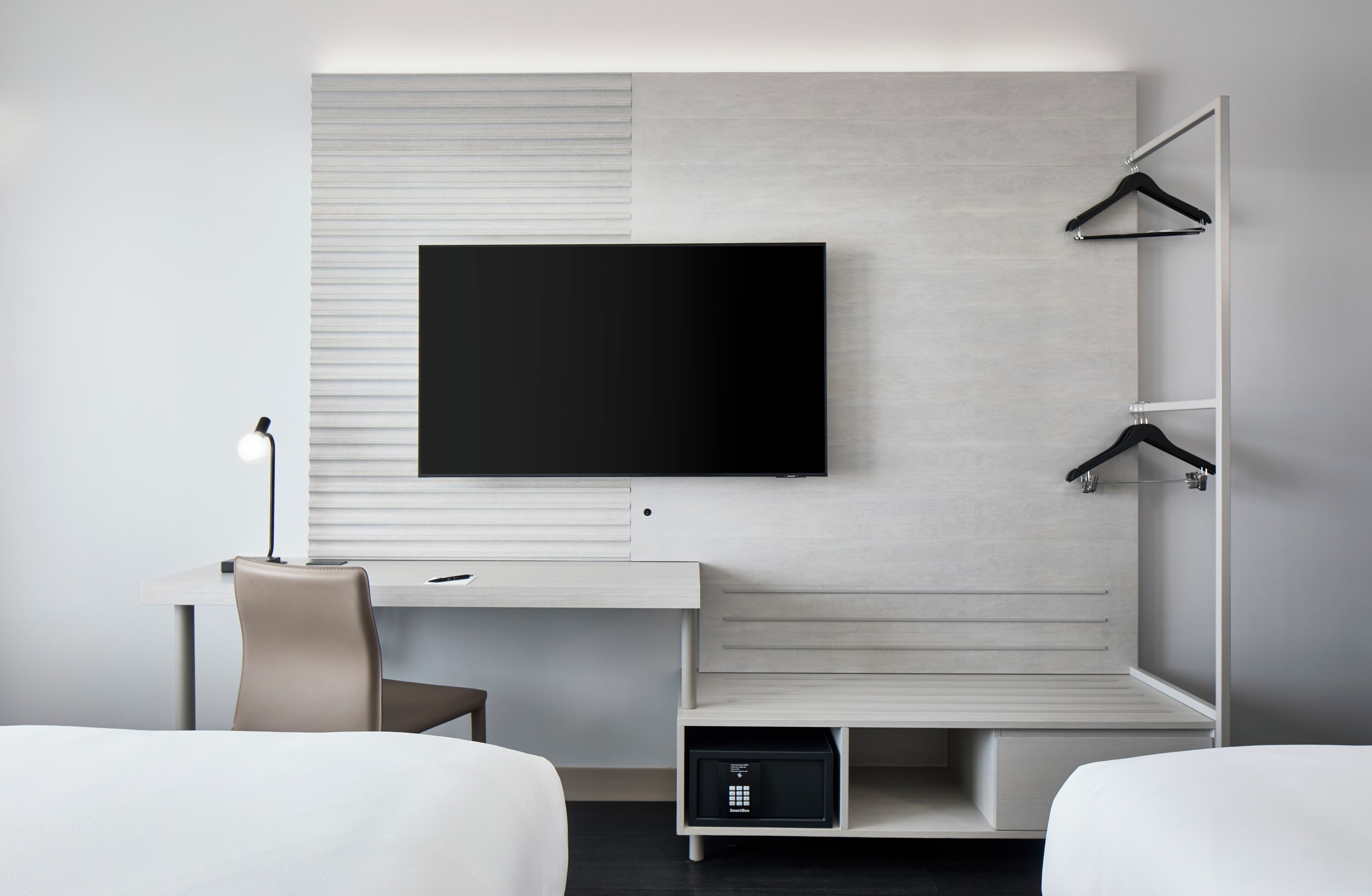 Een moderne hotelkamer met een flatscreen televisie aan de muur, een bureau met een lamp, een stoel en een garderobe met daarin hangende kleding.