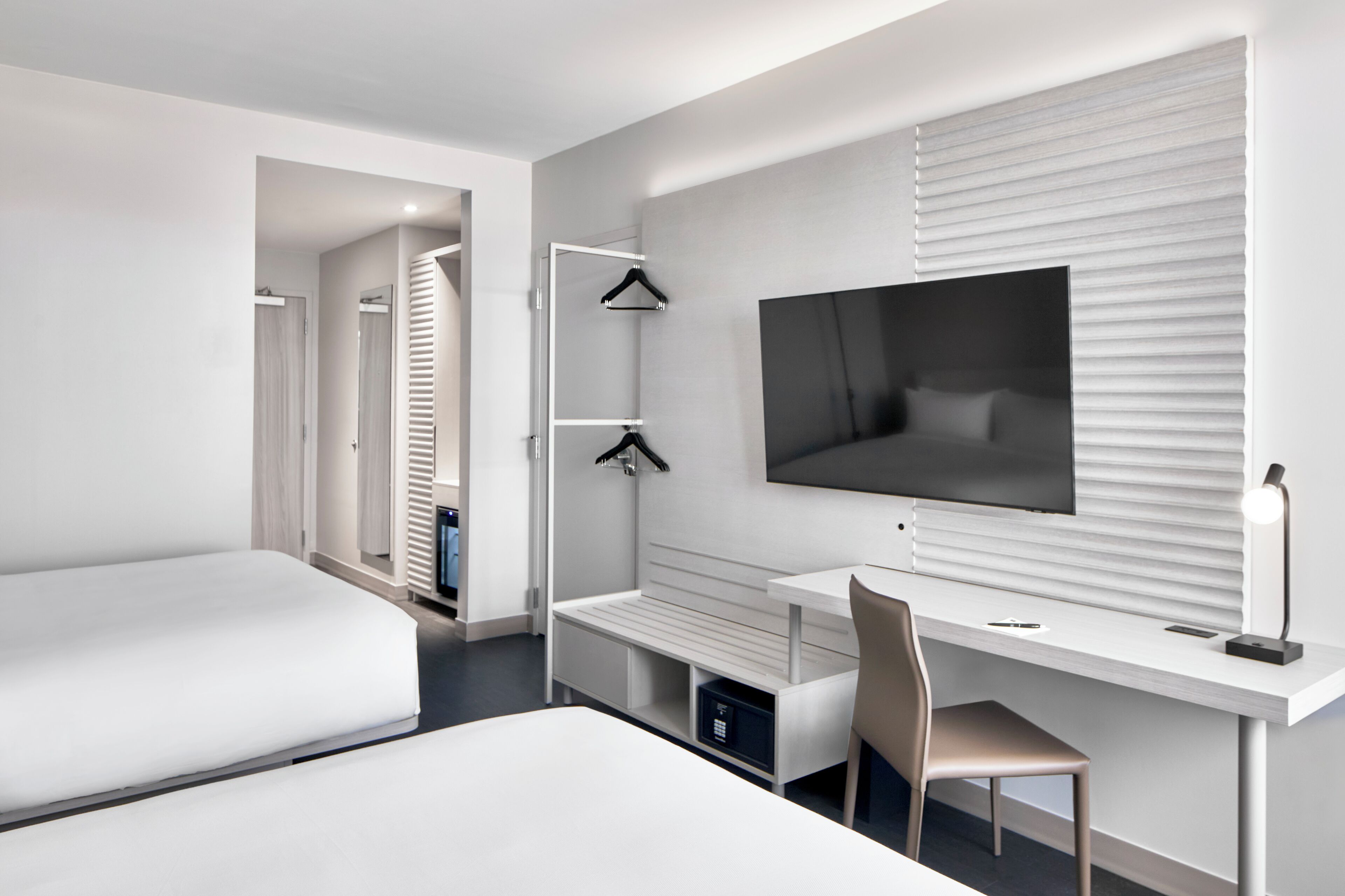Ein modernes Hotelzimmer mit einem großen Bett, einem Schreibtisch mit Stuhl, einem an der Wand montierten Fernseher und einem Schrank mit Spiegel.