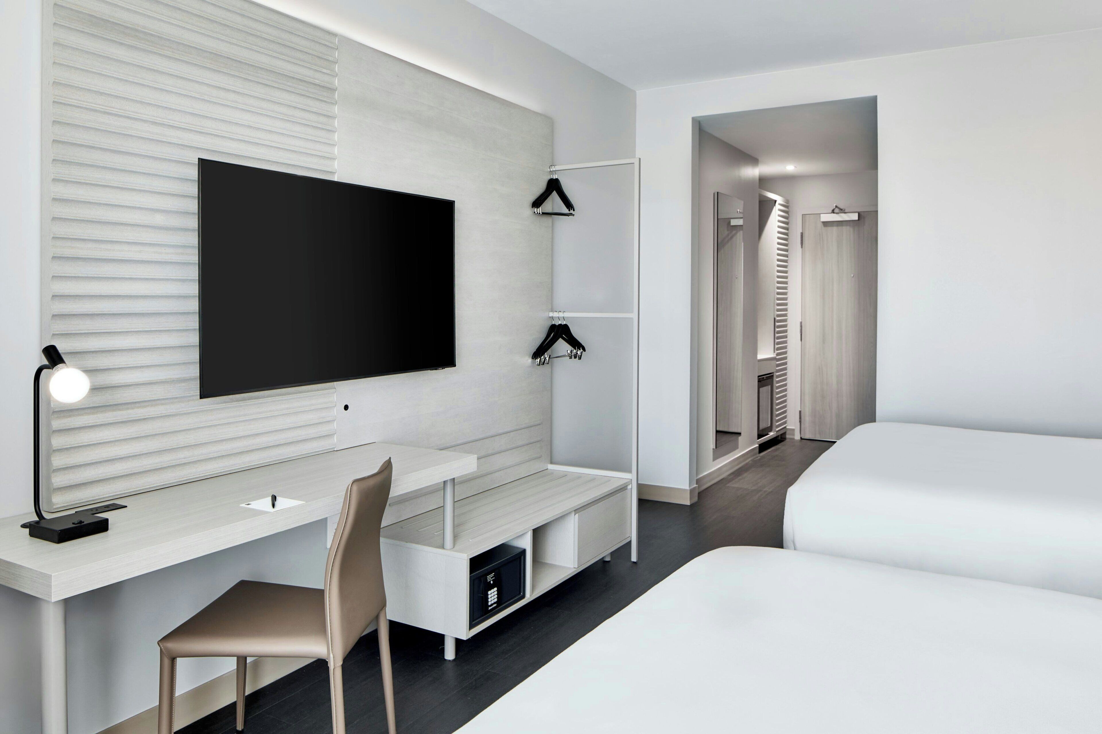 Una habitación de hotel moderna con una televisión de pantalla plana grande montada en la pared, un escritorio con una silla, una cama y un vestidor con espejo.