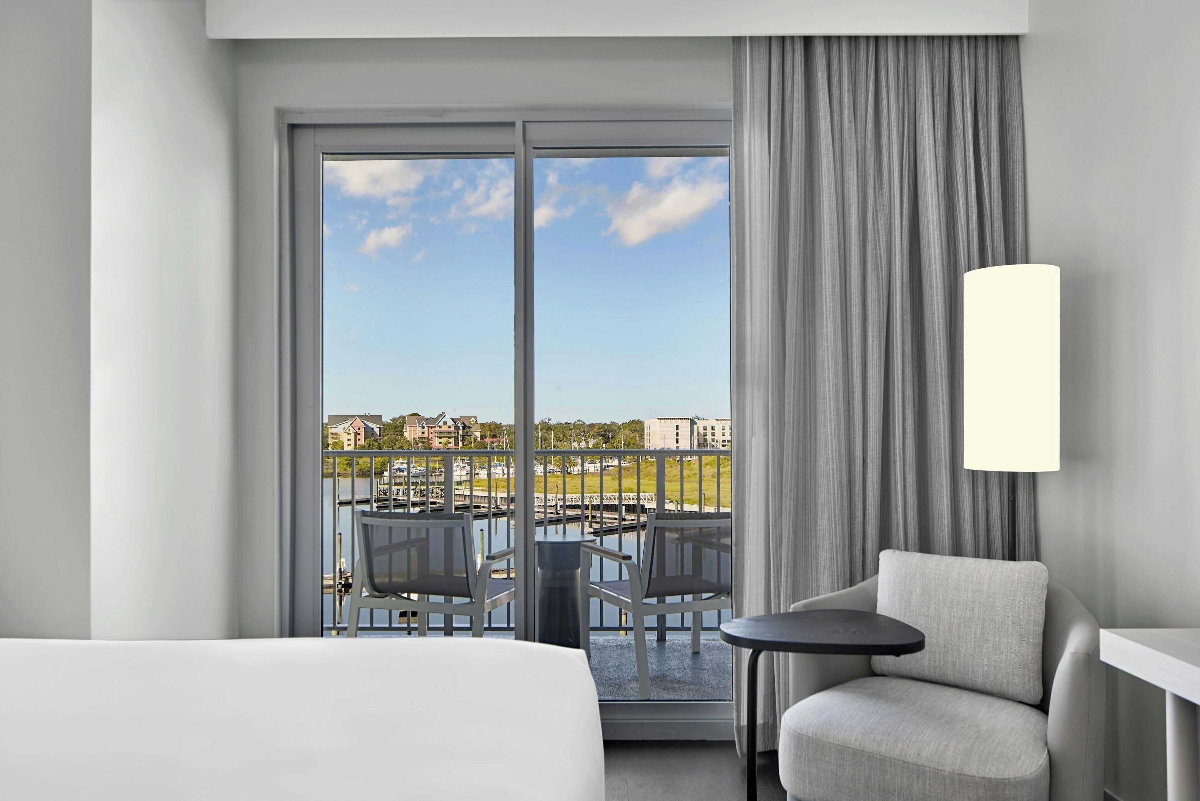 Ein Hotelzimmer mit großem Fenster, einem Balkon mit Stühlen, einem kleinen Tisch und einer Lampe.