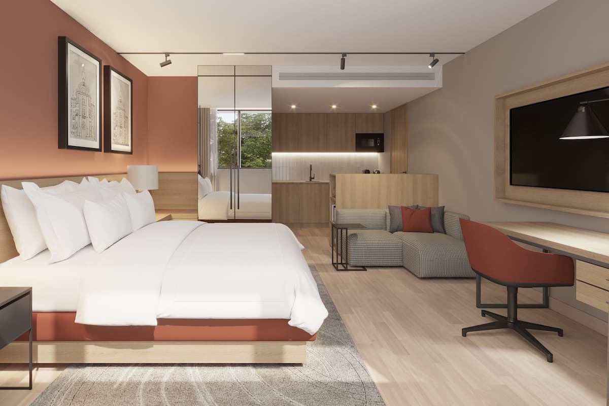A modern hotel room with a large bed, a desk, a chair, a TV, and a kitchenette.