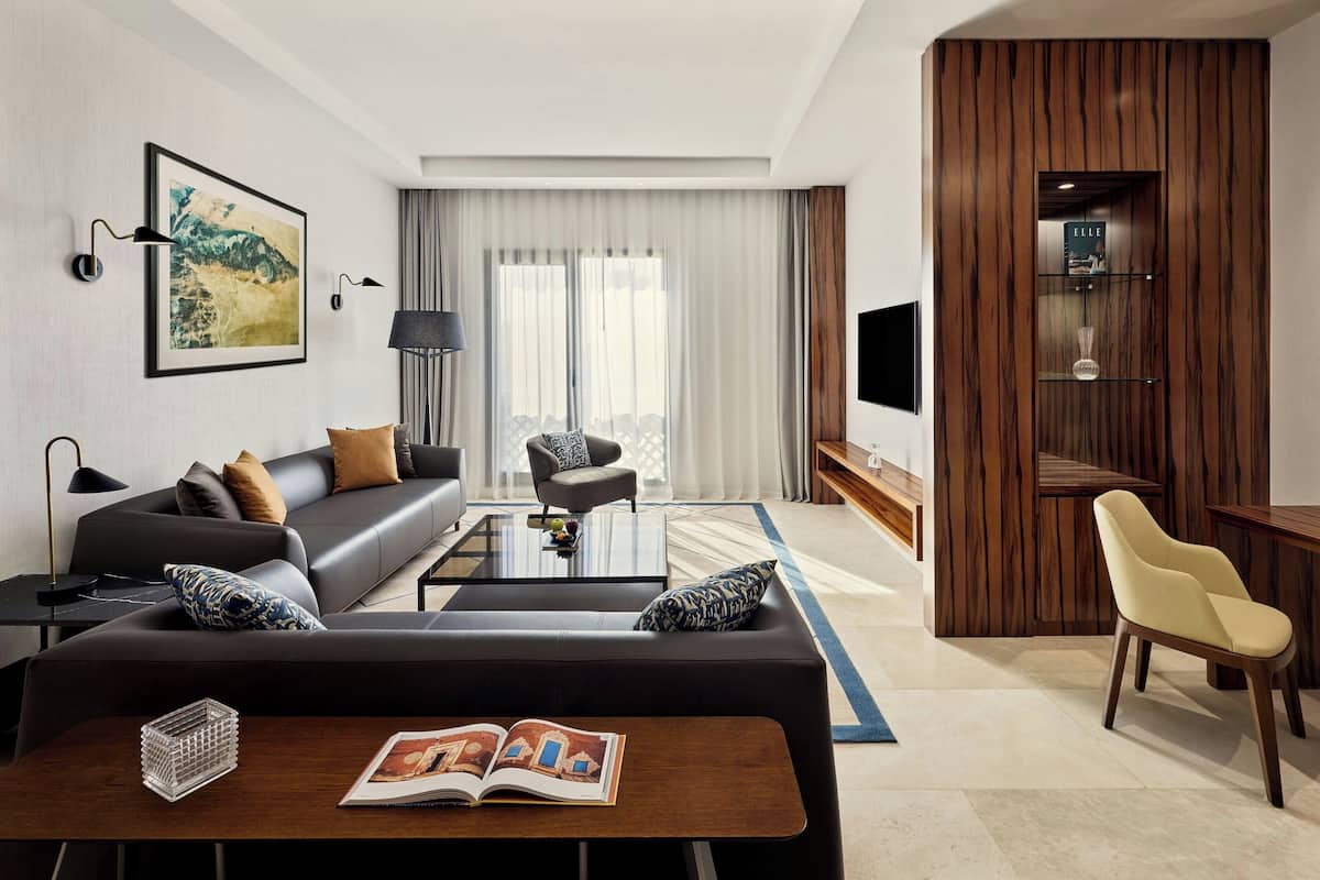 Una habitación de hotel moderna con un sofá de cuero oscuro, una mesa de centro de vidrio, un aparador de madera y un comedor con una silla amarilla.