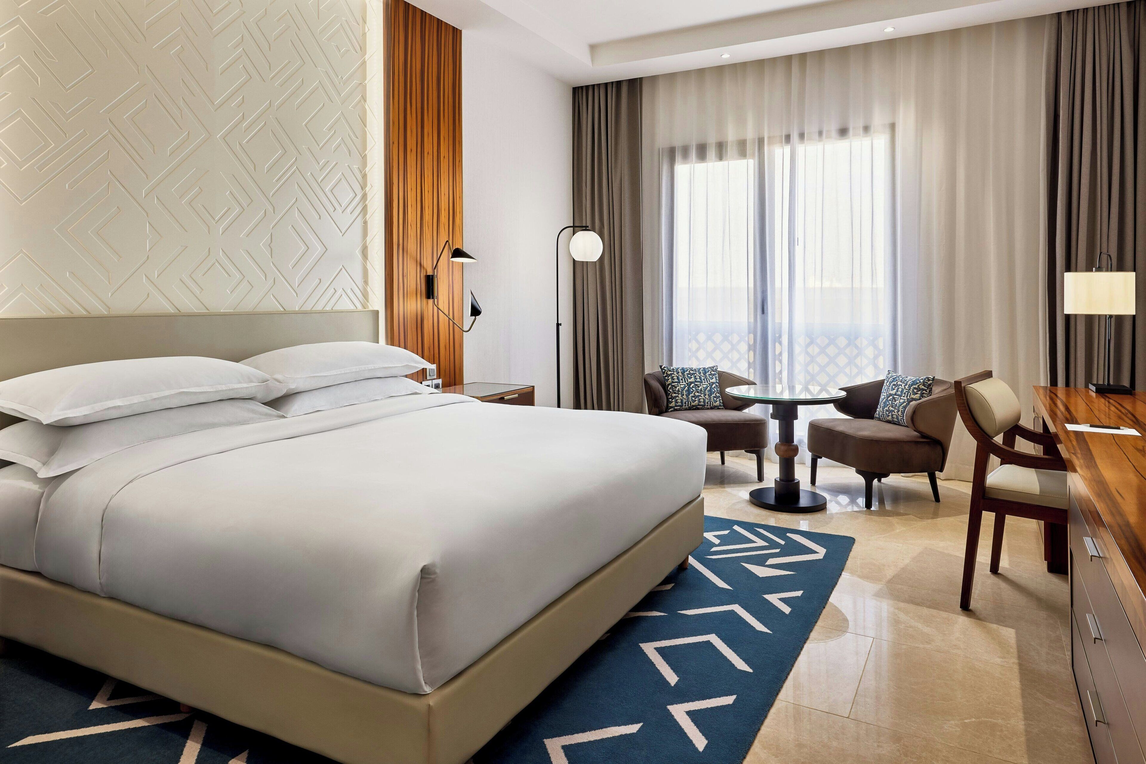 A hotel room with a large bed, a desk, a chair, a small table, and a view of the outdoors through the window.