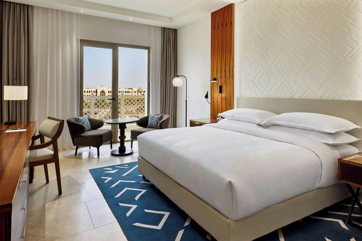 Habitación de hotel con una cama grande, un escritorio con silla, una mesita y vistas a la ciudad a través de amplios ventanales.