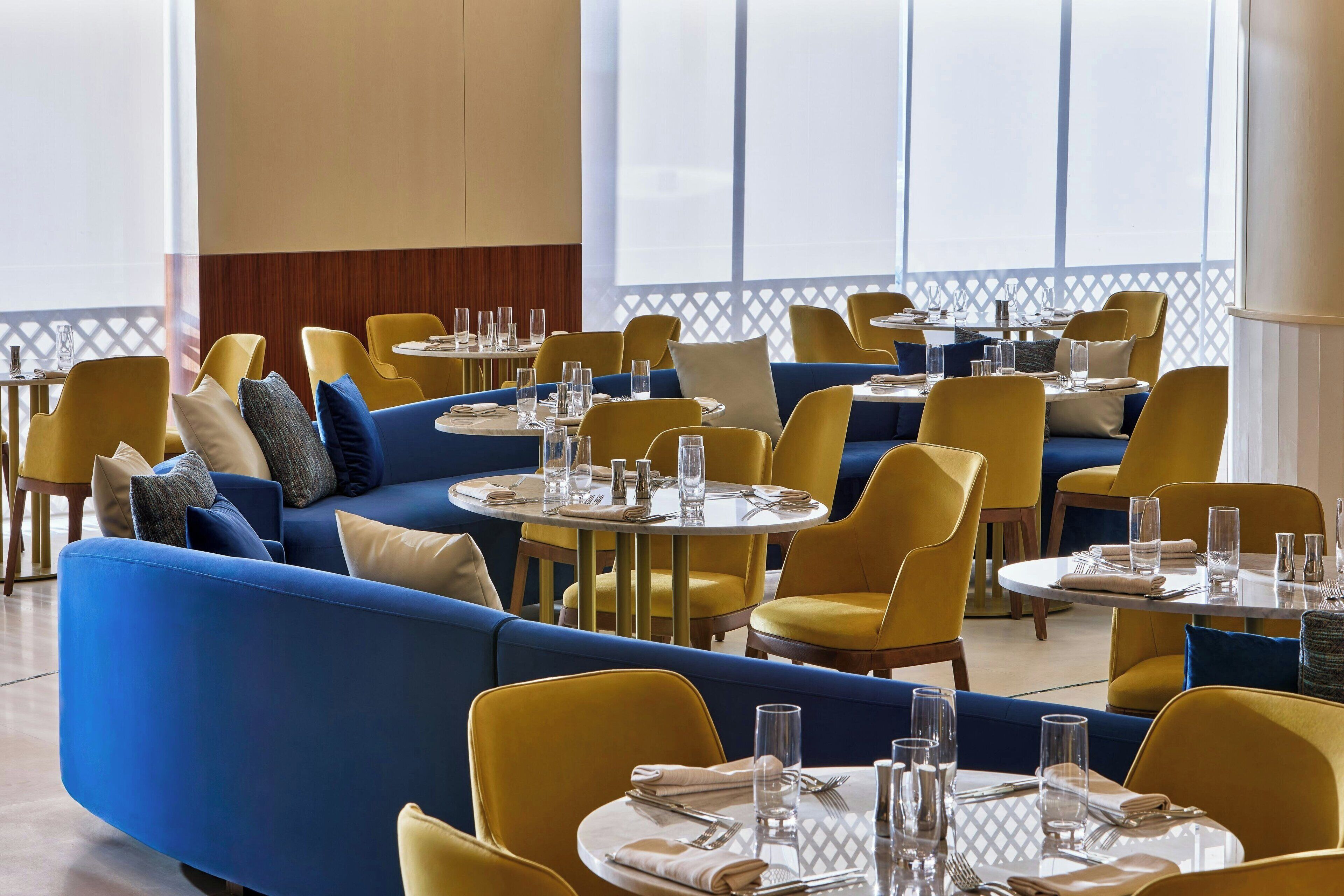 A restaurant with yellow and blue seating, round tables, and large windows.