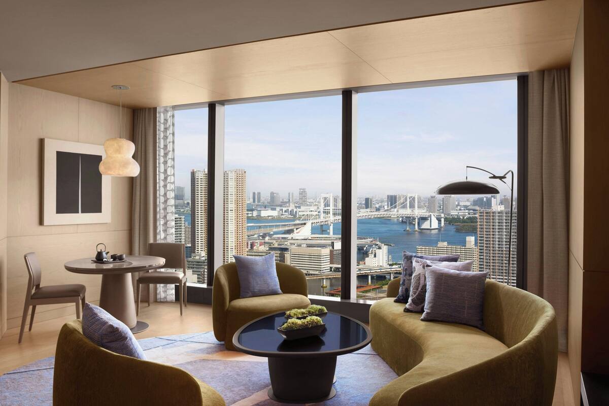 A modern hotel room with a large window offering a city view, a round coffee table with a plant, and a seating area with two sofas and a chair.