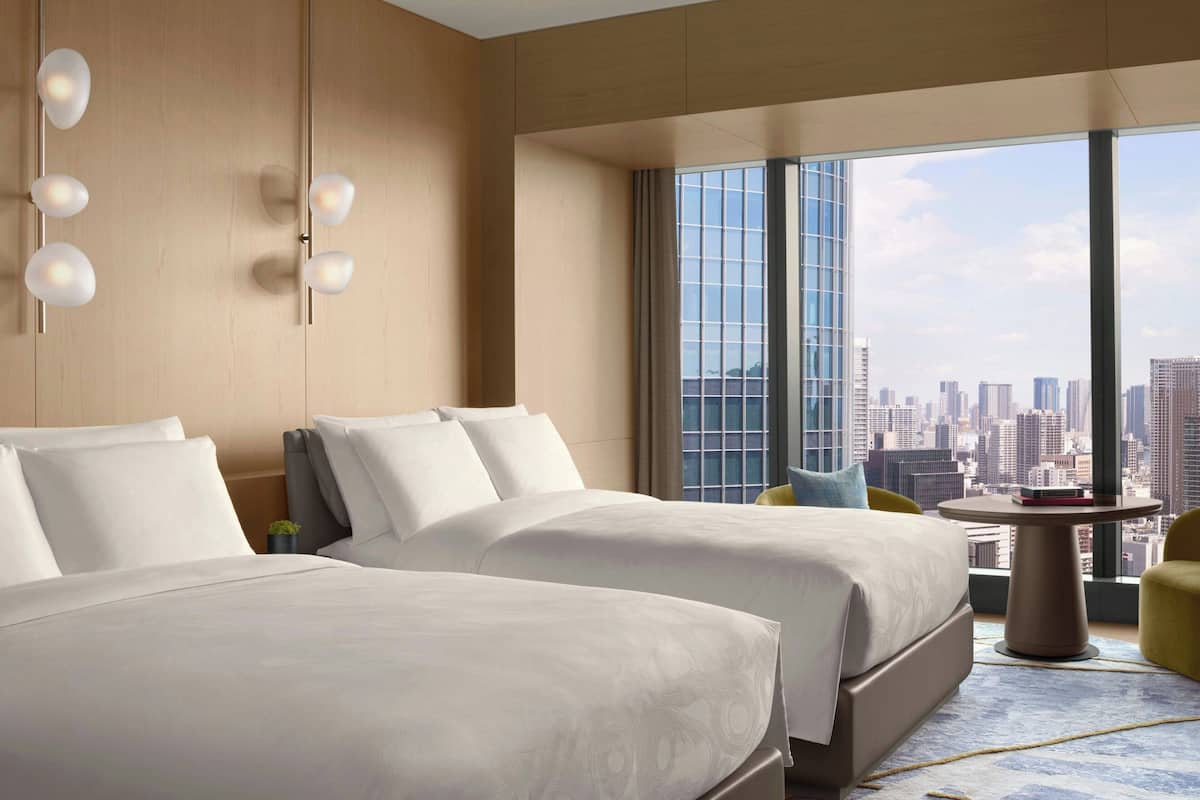 A modern hotel room with two beds, a city view, and a small table.