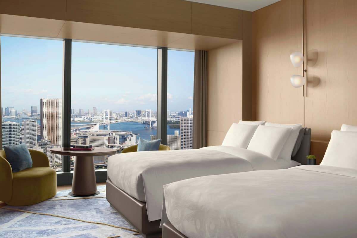 A hotel room with two beds, a view of a cityscape, a small round table, and a yellow armchair.