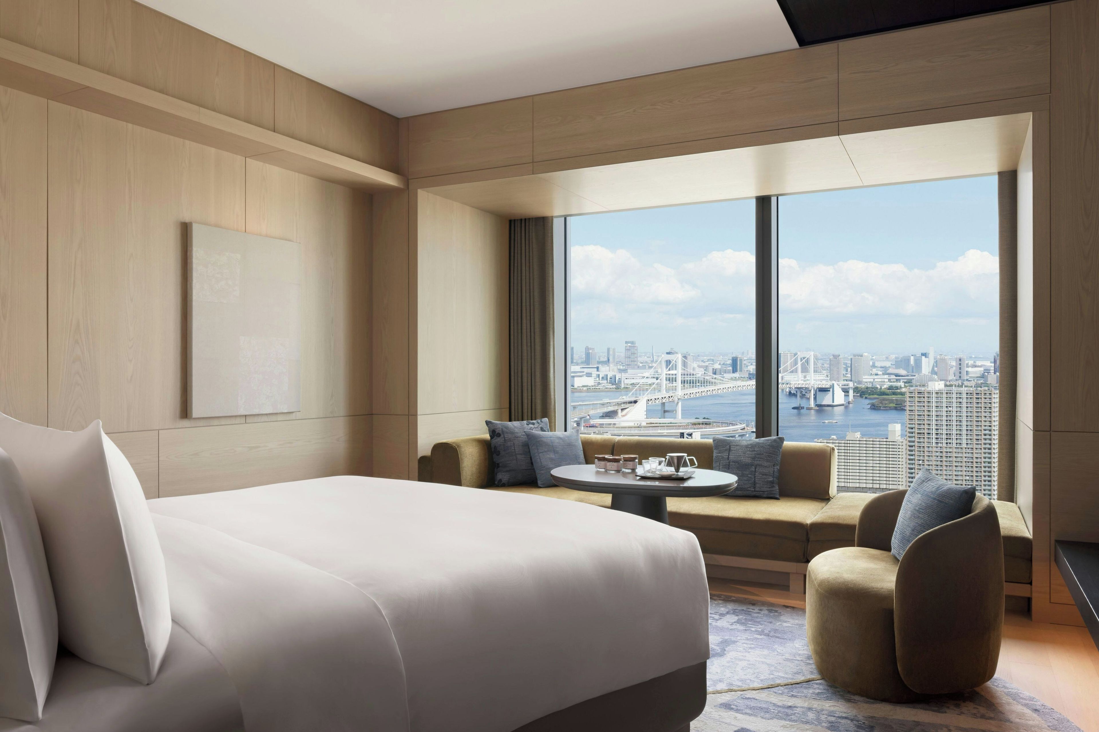 A modern hotel room with a large bed, a sofa, a coffee table, and a view of a cityscape through large windows.