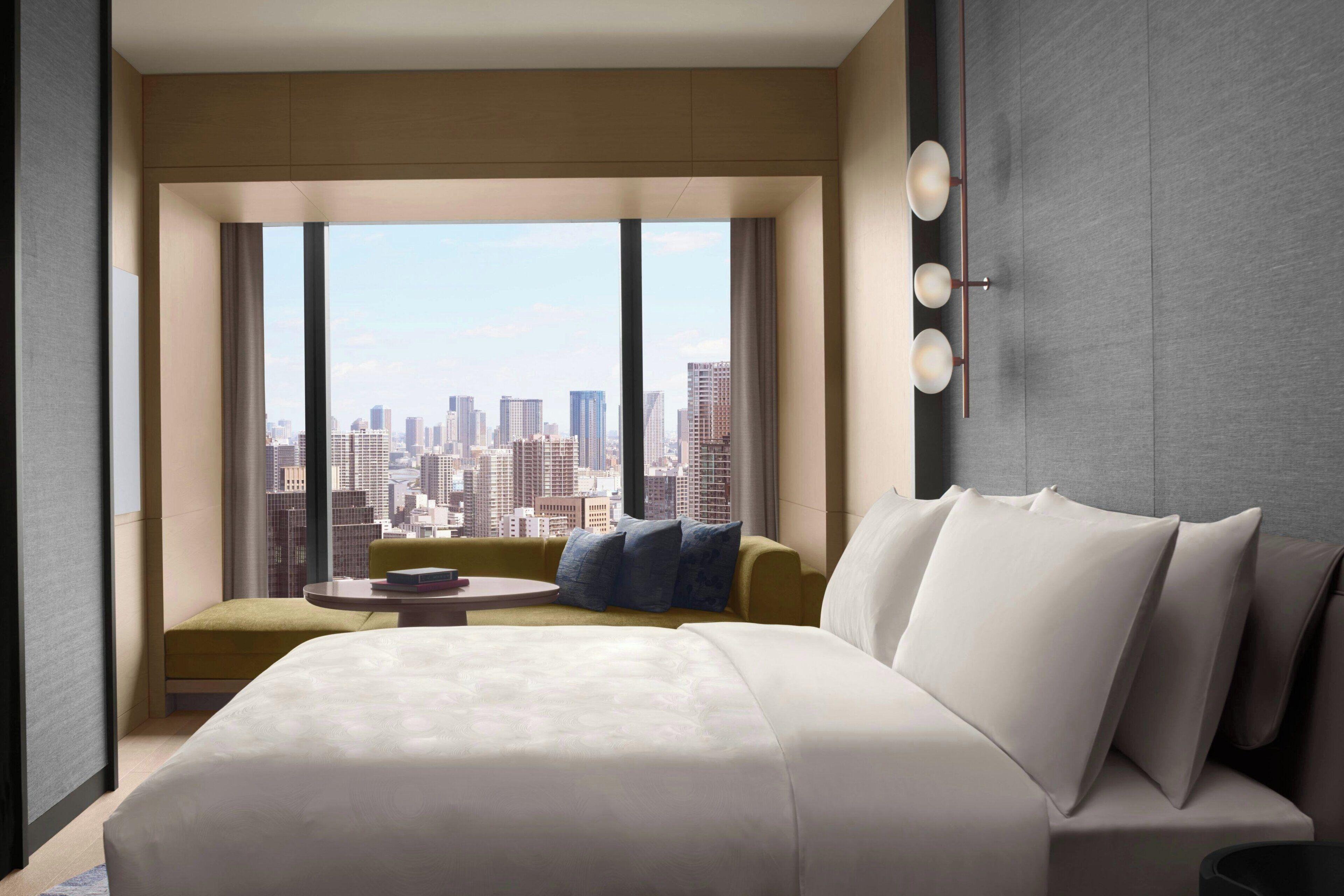 Une chambre d’hôtel moderne avec un grand lit, un canapé vert, une petite table ronde et une vue sur la ville à travers de grandes fenêtres.