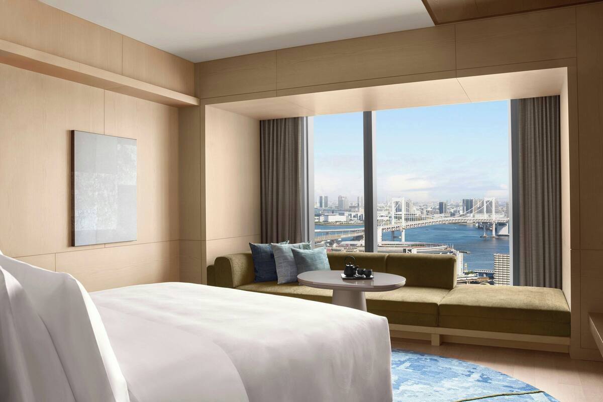 A hotel room with a large bed, a sofa, a small table, and a view of a cityscape and water.