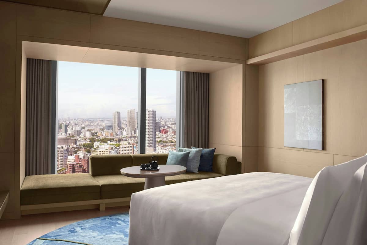 A hotel room with a large bed, a sofa, a round table, and a city view through large windows.