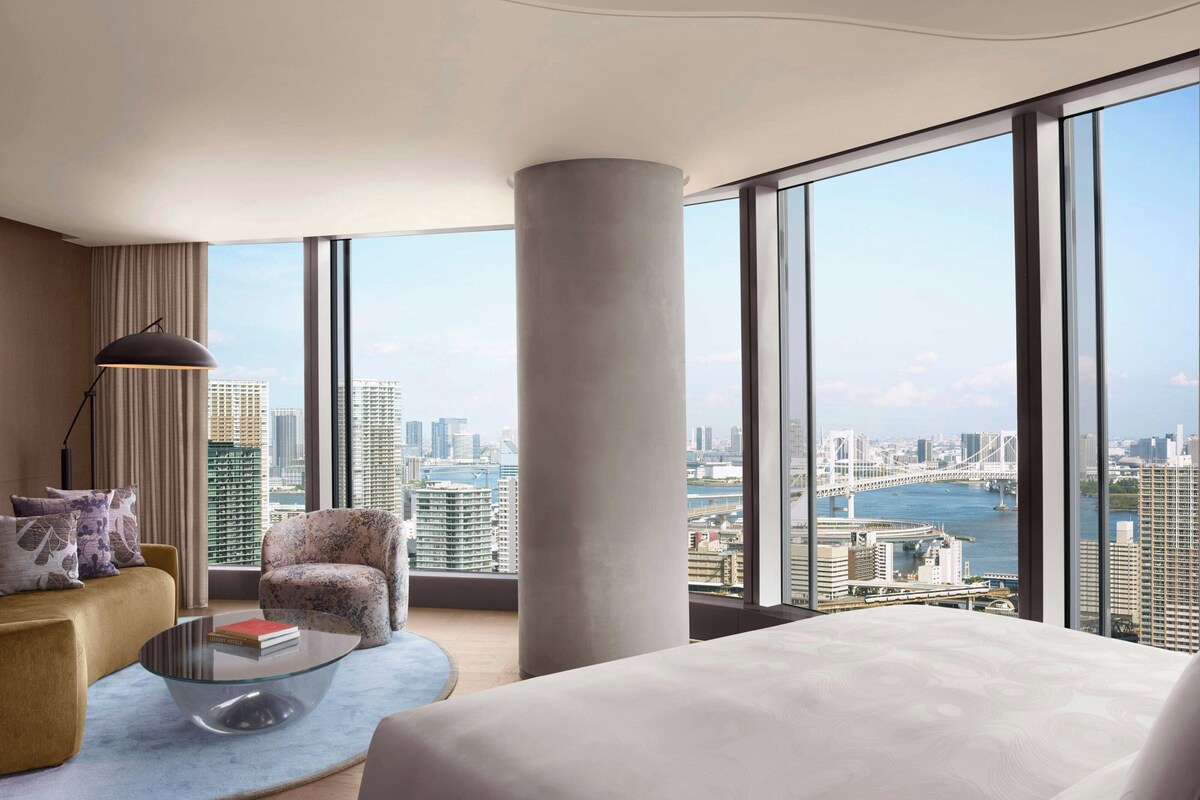 A modern hotel room with a large window offering a city view, a comfortable sofa, a glass coffee table, and a large bed.