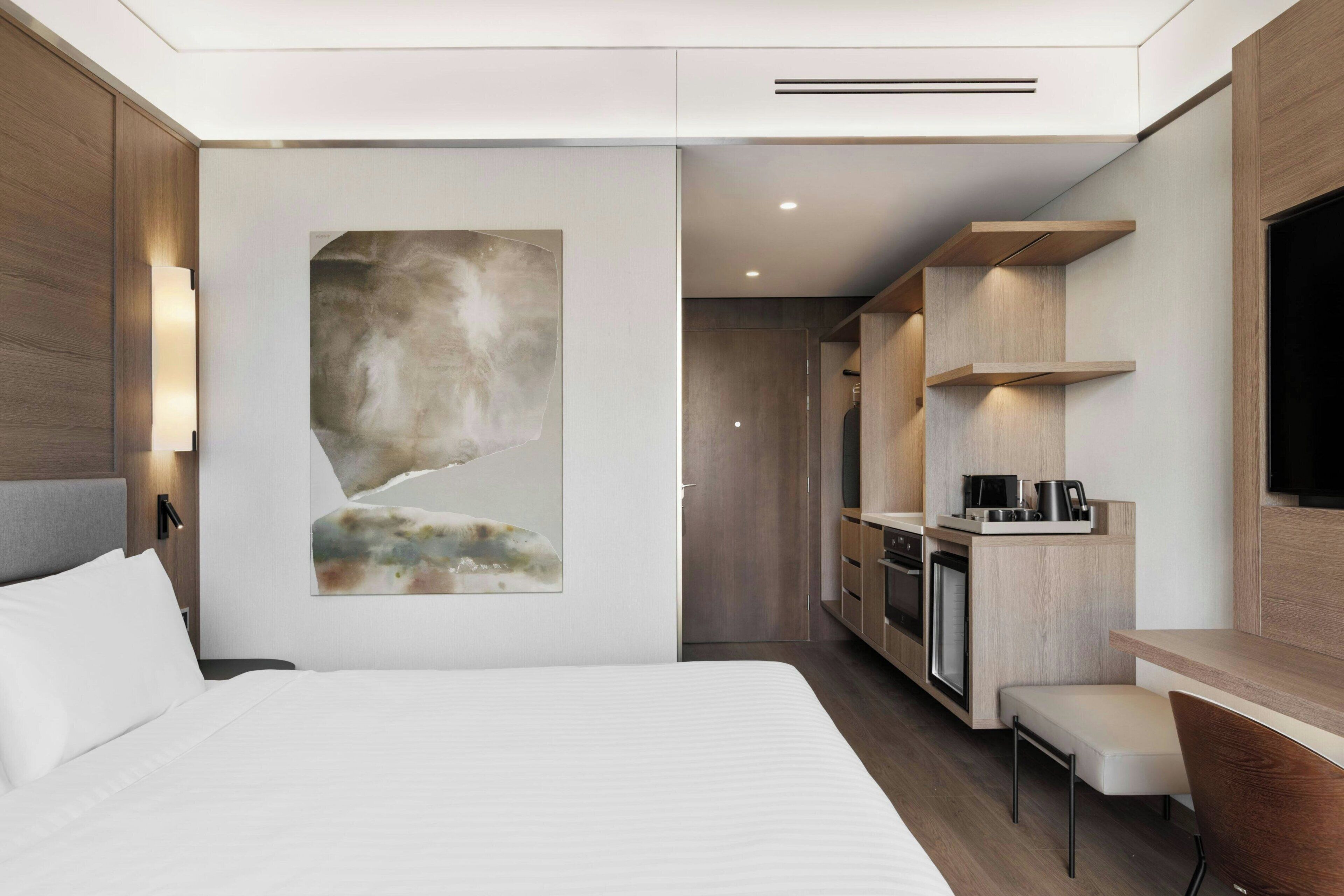 Une chambre d’hôtel moderne dotée d’un grand lit, d’une petite kitchenette, d’un coin salon et d’un grand tableau abstrait accroché au mur.