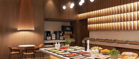 A modern restaurant interior with a central counter displaying an assortment of food items, a seating area with chairs, and a wall-mounted lighting fixture.