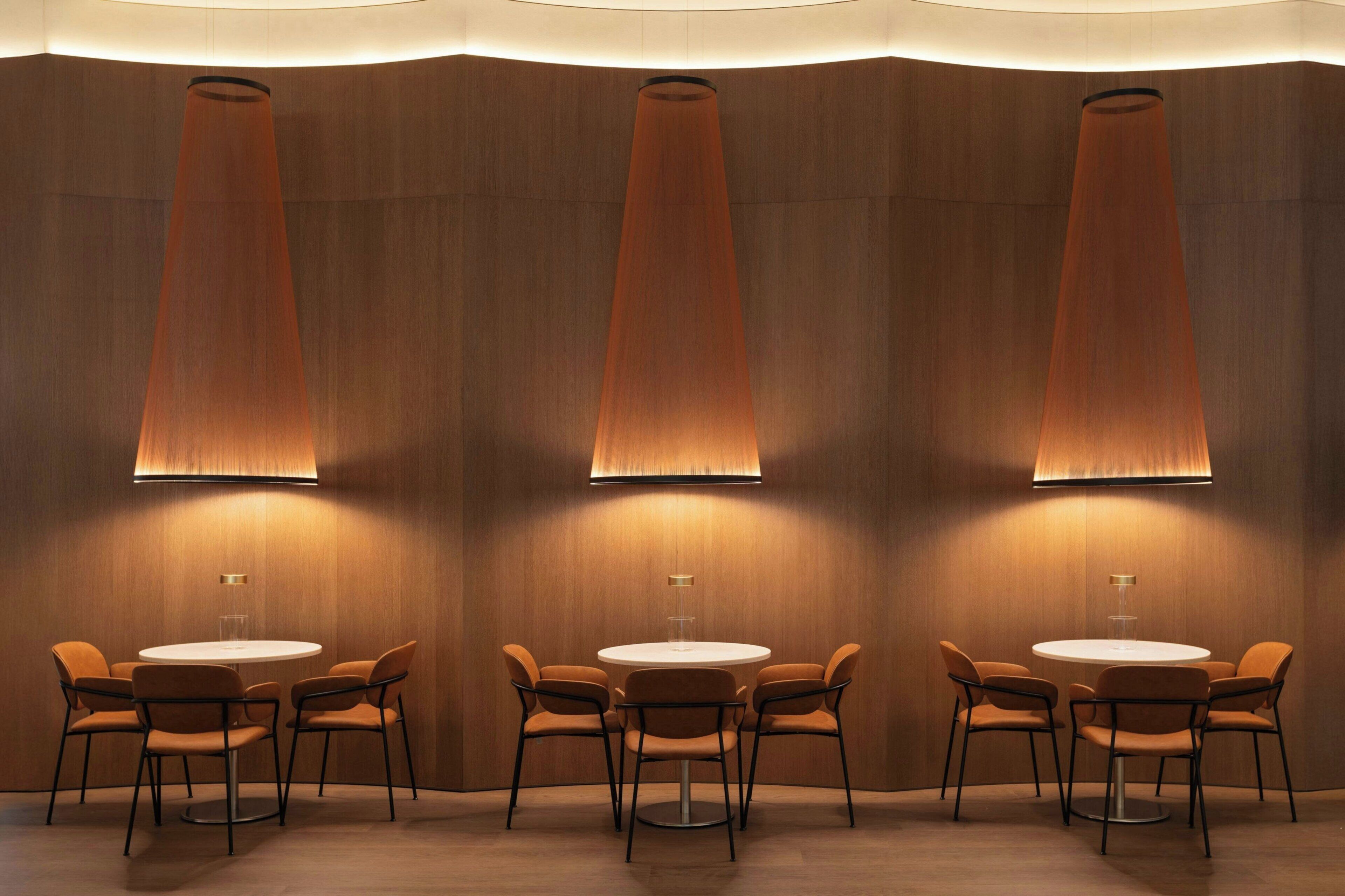 A modern dining area with round tables and orange chairs, illuminated by tall, conical lamps.