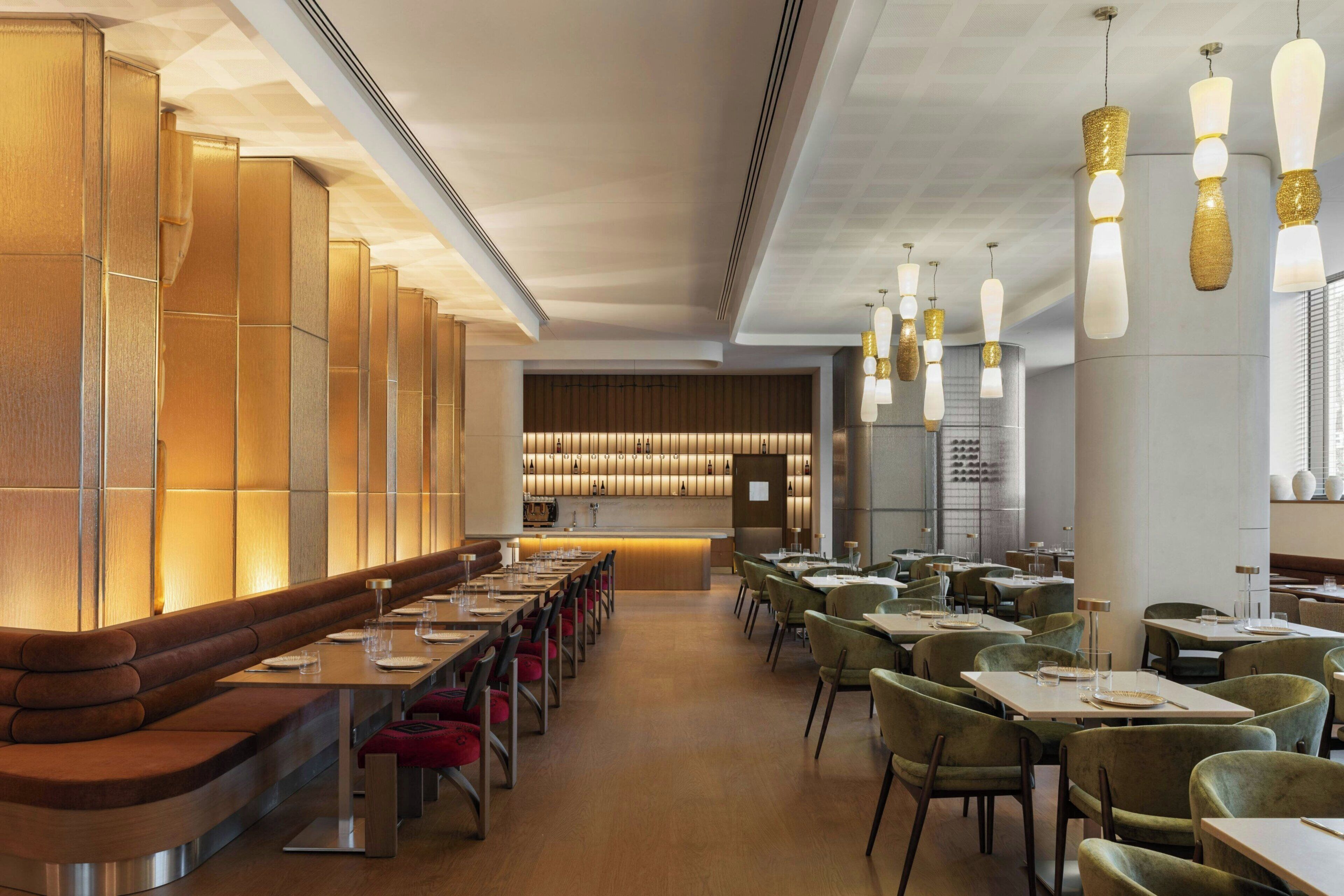 A modern restaurant with long communal tables, cushioned seating, and pendant lighting.