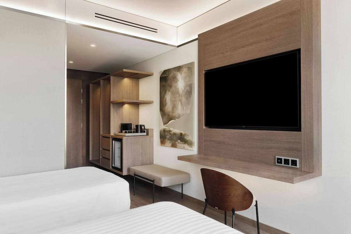 Ett modernt hotellrum med en stor platt-TV som är monterad på väggen, en säng med ett vitt täcke, en träbänk och ett litet skrivbord med en kaffebryggare.