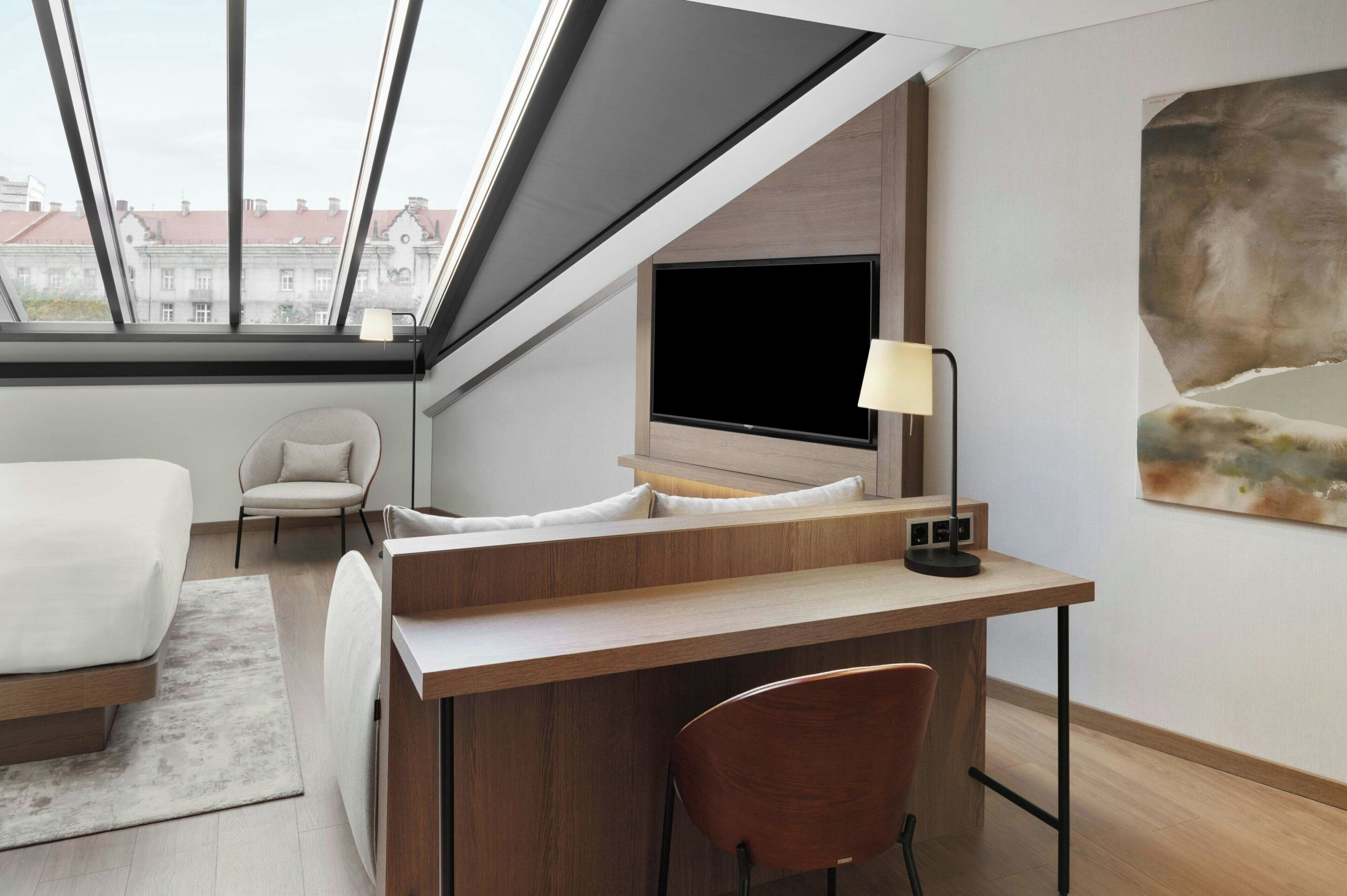 A modern hotel room with a large skylight, a bed, a desk, a chair, and a painting on the wall.