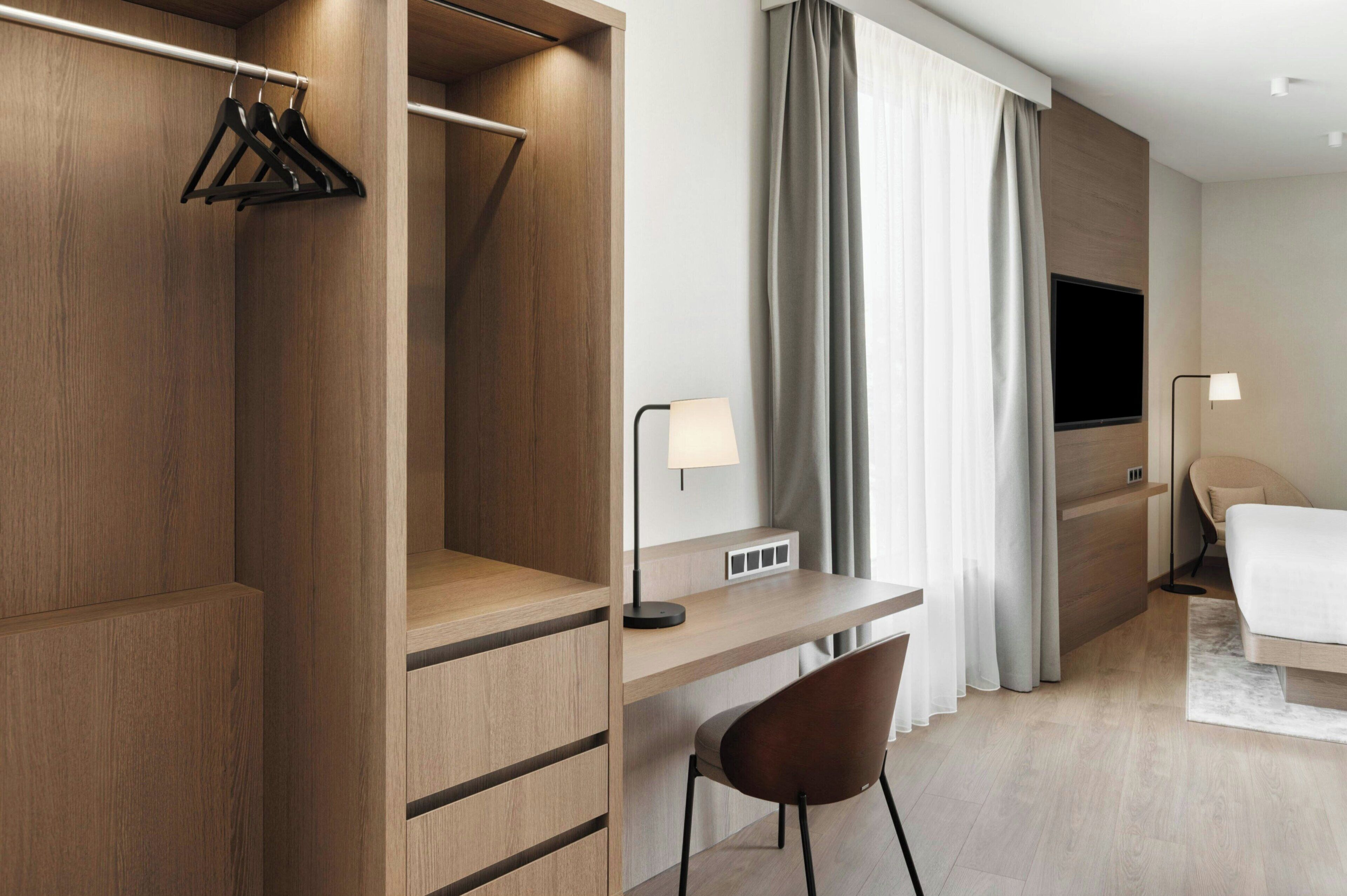 Quarto de hotel moderno com guarda-roupa de madeira, uma escrivaninha com luminária, uma cama e uma televisão.
