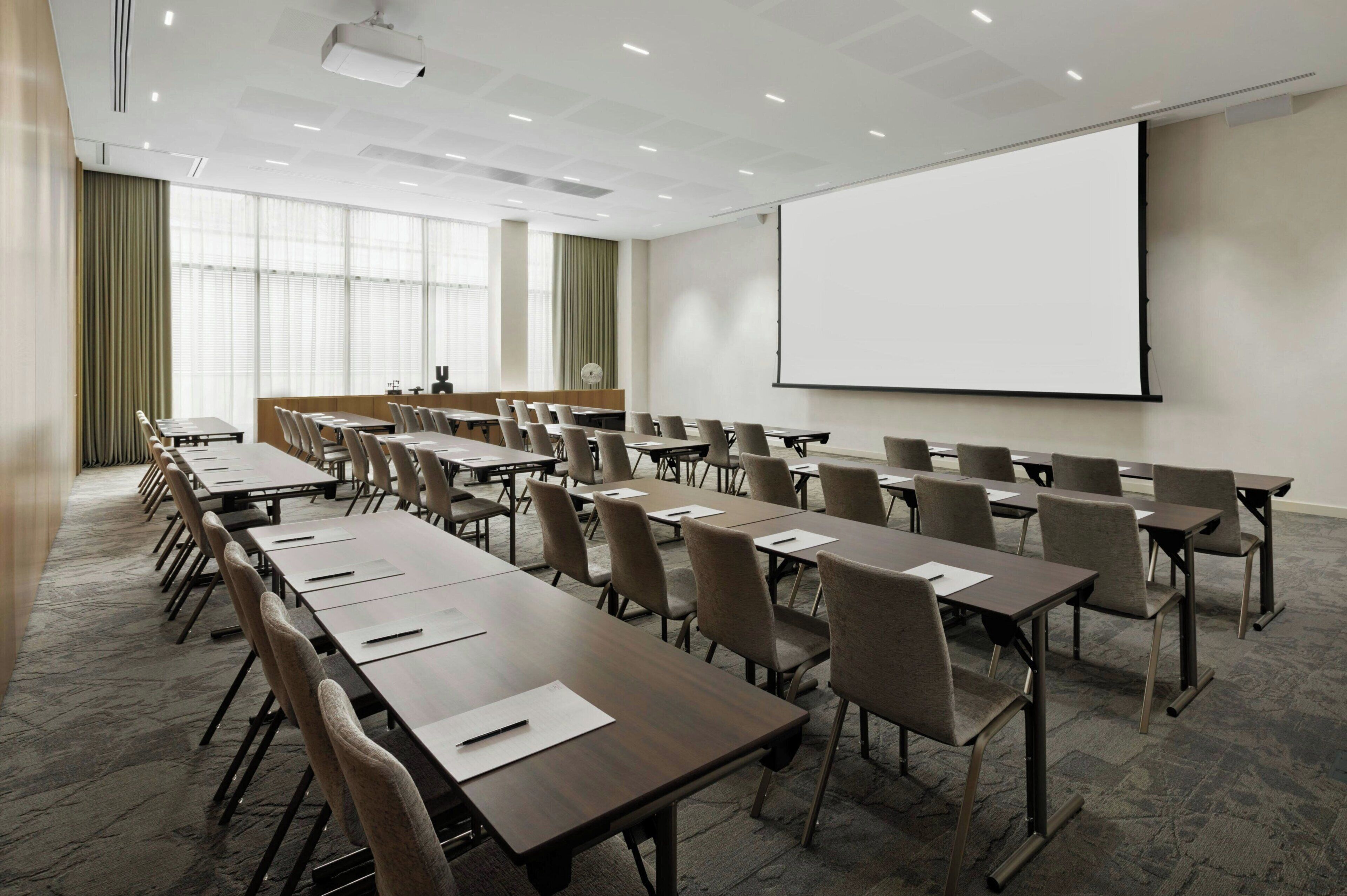 Un salón de conferencias con largas mesas y sillas, una pantalla de proyector y un ventanal con cortinas.
