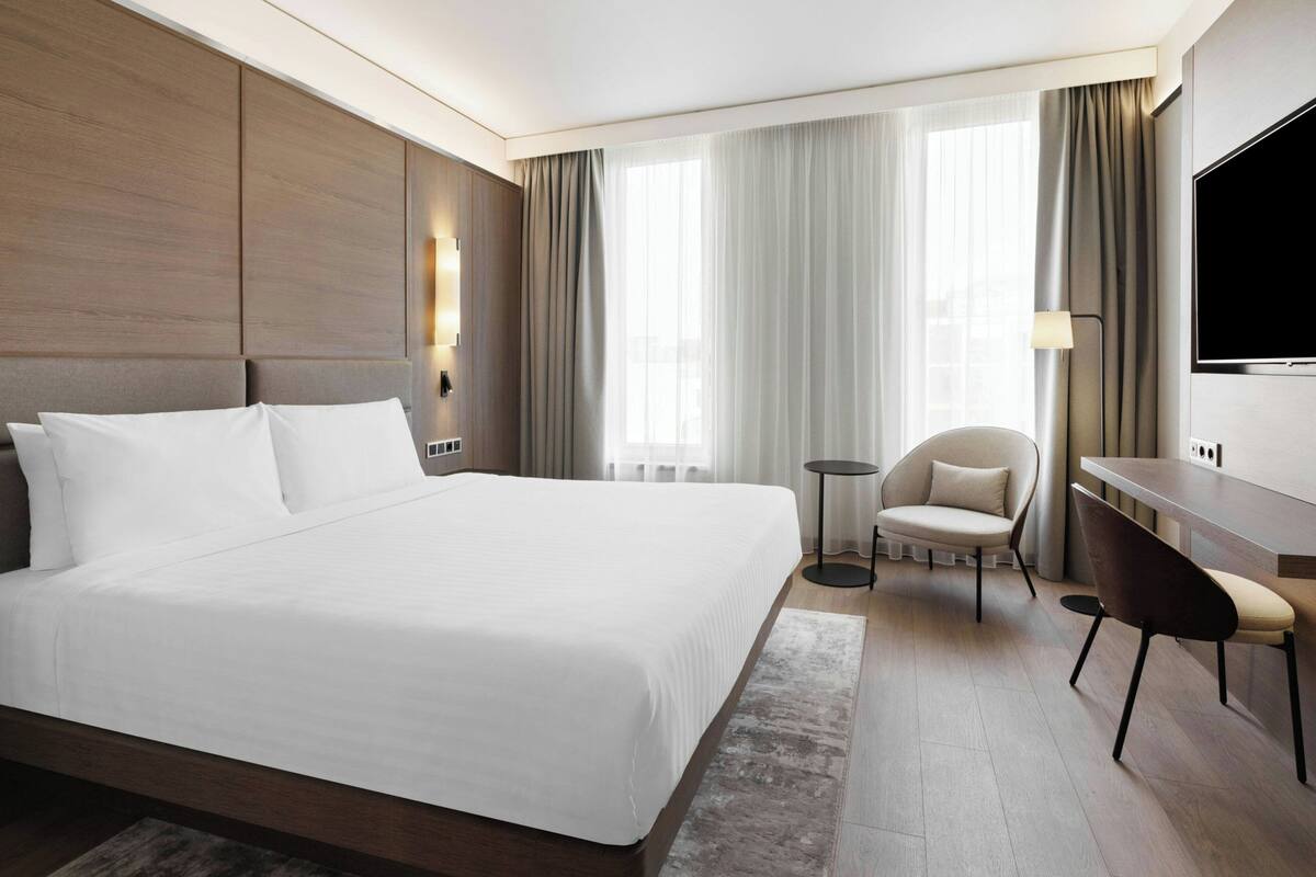 Ett hotellrum med en stor säng, ett skrivbord, en stol, en tv och ett fönster med gardiner.