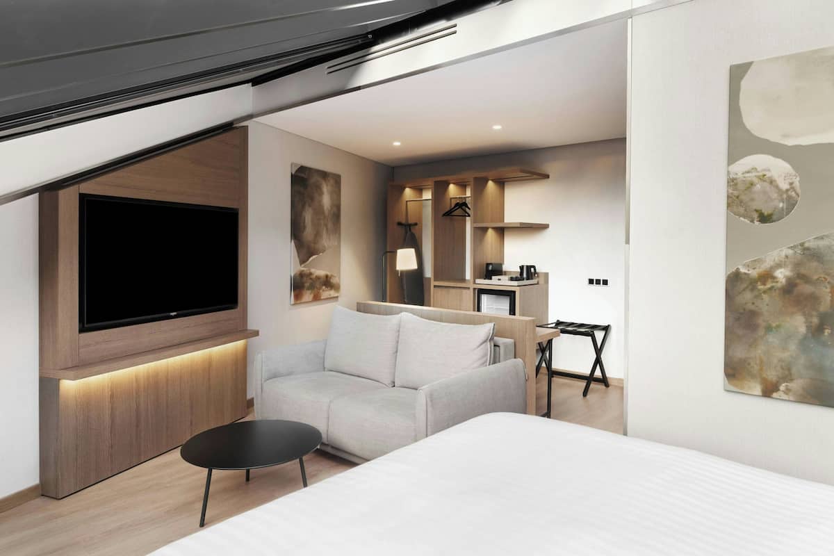 Ett modernt hotellrum med en stor platt-TV, en soffa, ett litet bord och en säng. Det finns också en liten köksdel med en kaffemaskin och en spegel.